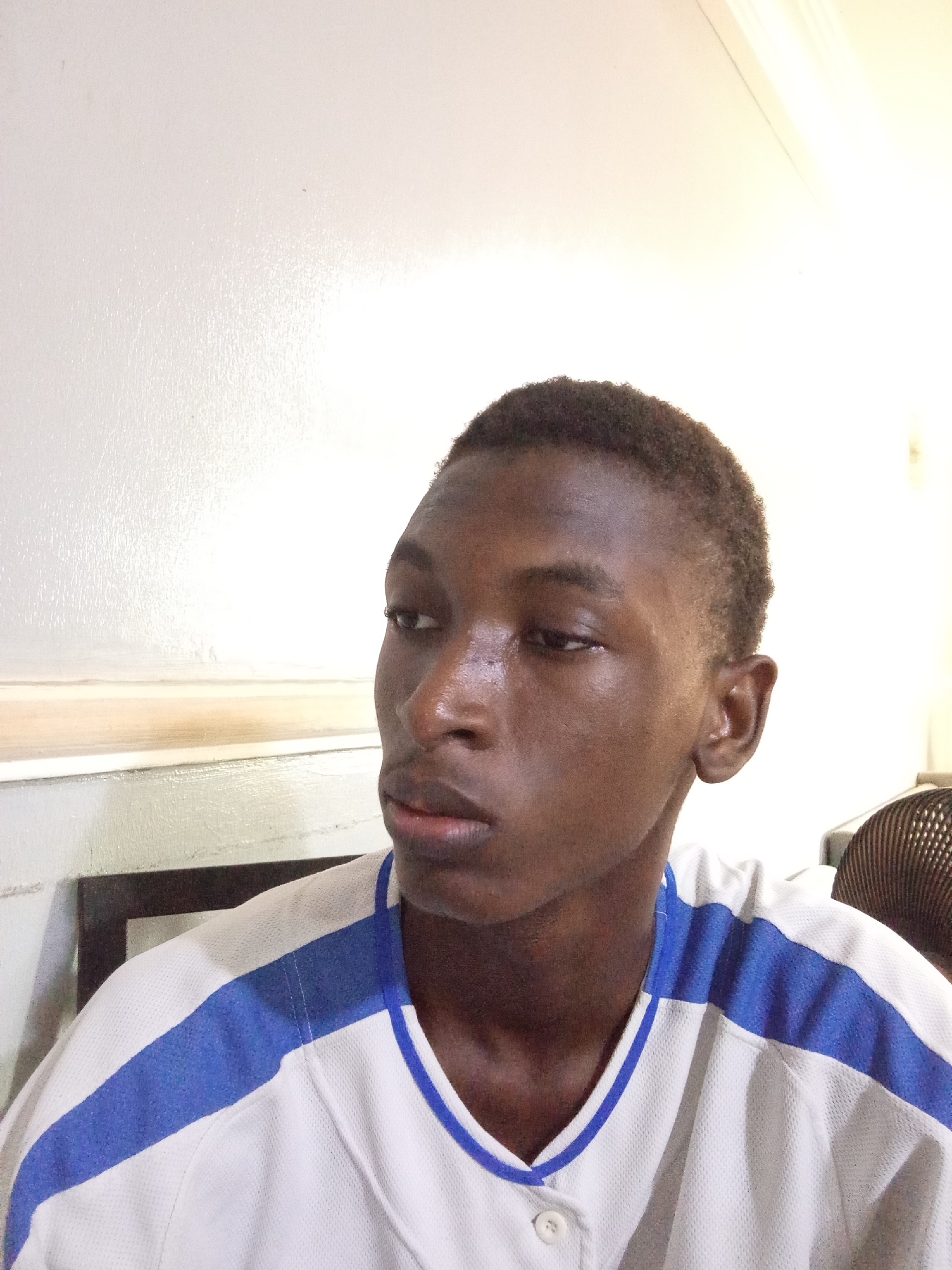 A medium shot of a young Black man with short, dark hair, wearing a white baseball-style jersey with blue shoulder stripes and a single button visible. The man is looking slightly off-camera to his left, with a serious or thoughtful expression. The background includes a white wall, part of a dark-framed object (possibly furniture or a window), and part of a black mesh object (possibly a chair). The location is stated to be in the city of Unknown, Unknown.