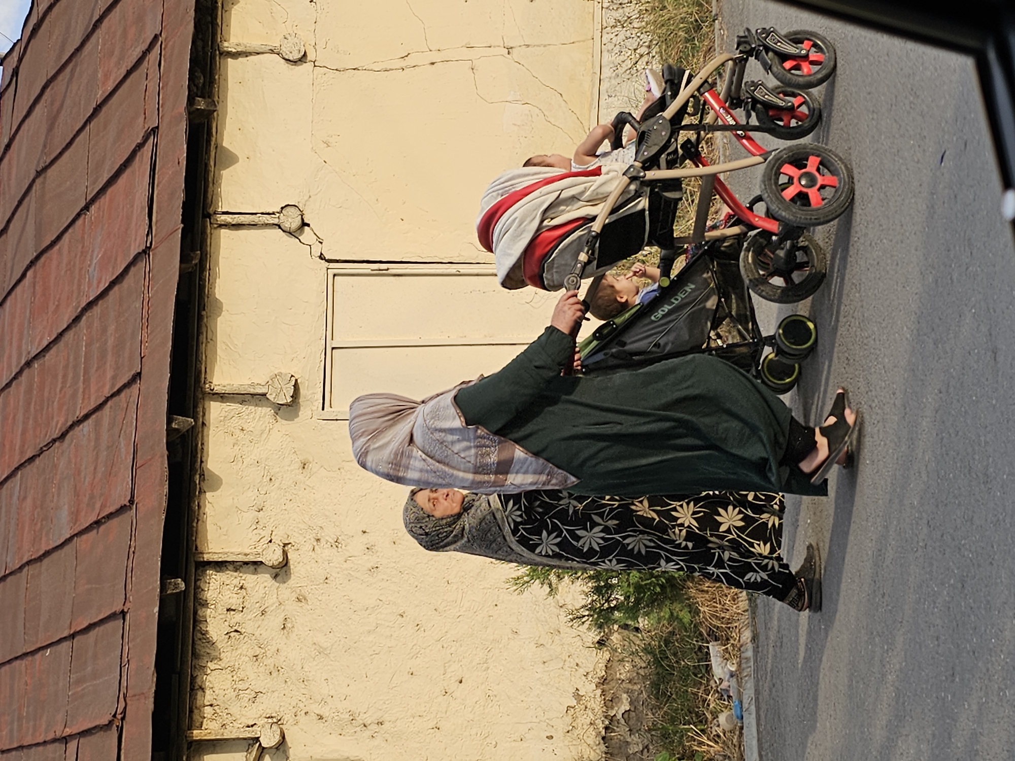 In Özgön, Kyrgyzstan, two women are walking on a paved road in front of a weathered, cream-colored building with a red corrugated metal roof. The woman on the left wears a black headscarf with a floral pattern and a matching black and white patterned dress. She is looking to her left, away from the camera. The woman on the right, closer to the camera, wears a light-colored headscarf with a patterned border and a dark green robe. She is pushing a double stroller. The stroller on the left is black, and the one on the right is beige with red accents. A young child, wearing a white top, is visible in the right stroller. Another child, with dark hair, is in the left stroller, holding his hand up to his face. The ground is dry asphalt with shadows cast from the people and stroller. Sparse vegetation and debris are visible along the edge of the road.
