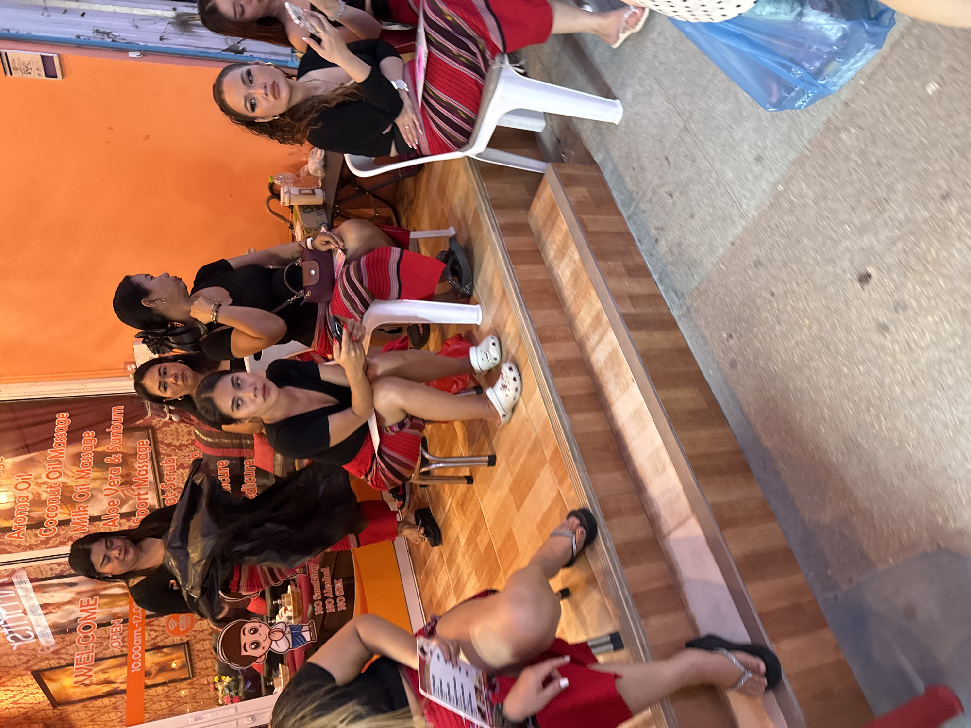 In Patong, Thailand, a group of women, some in uniform red and striped skirts, are seated or standing in what appears to be a massage parlor. A sign in the background lists services like "Aroma Oil," "Coconut Oil Massage," and "Milk Oil Massage." One woman in the foreground is holding a brochure, while another is looking at her phone. The floor is tiled, and there are wooden steps leading up to the area where the women are situated. A blue plastic bag is visible on the ground in the lower right of the frame.