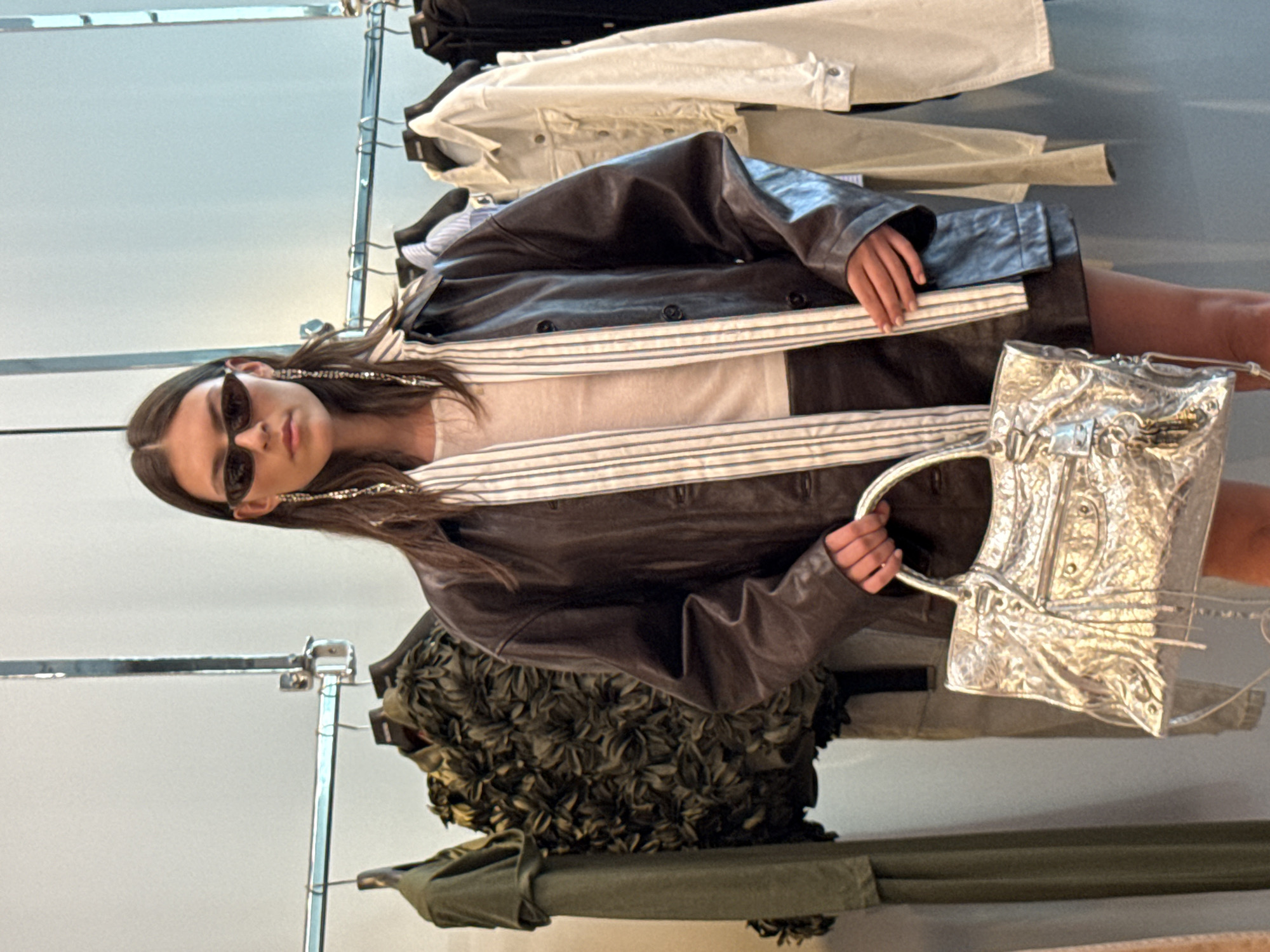 A young woman stands in front of a clothing rack in what appears to be a retail store. She is wearing a dark leather jacket over a white top with thin blue stripes, sunglasses, and long dangling earrings. She is holding a shiny silver handbag. The background shows various articles of clothing hanging on the rack. The image appears to be taken indoors in Dubai, United Arab Emirates.