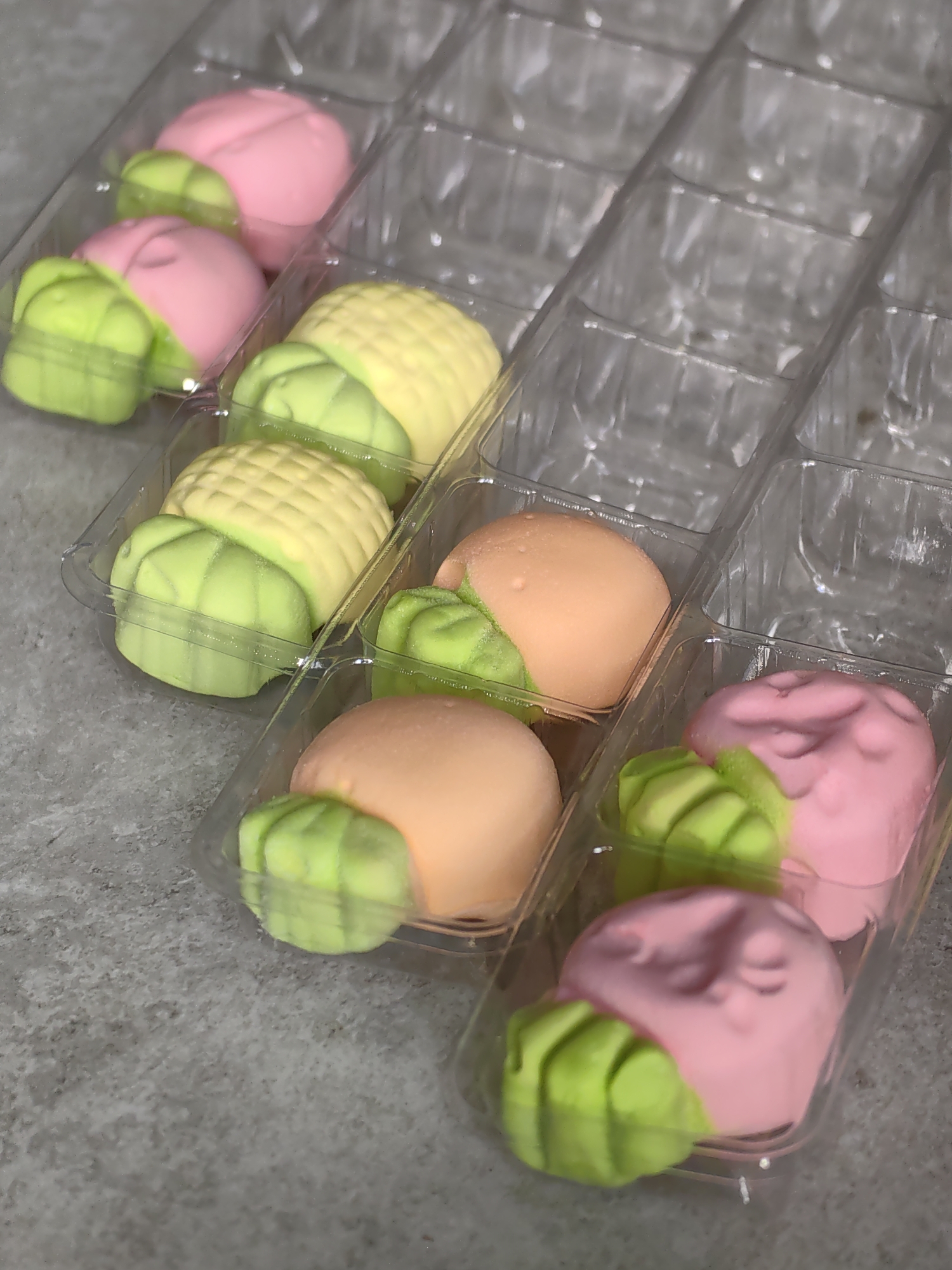 In the city of Fanipal, Belarus, a close-up, high-angle shot captures a plastic container holding individually packaged mochi. The mochi are shaped to resemble fruits and vegetables. In the top row, two pink mochi are arranged, each with green, leaf-like accents. To their right, a yellow mochi, resembling corn on the cob, is visible. The second row features two green mochi with yellow tops, resembling corn, followed by a peach-colored mochi with green accents, possibly a plum or apricot. The bottom row shows a peach-colored mochi with green accents, and two pink mochi with green accents. The clear plastic packaging has multiple individual compartments, suggesting a retail display. The mochi are placed on a textured gray surface. The overall scene is well-lit, highlighting the vibrant colors and detailed craftsmanship of the confectionery.