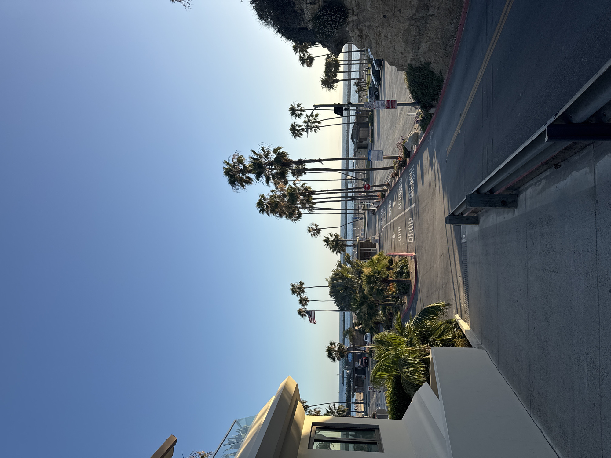 This image depicts a coastal scene in Newport Beach, California, USA. It captures a view from an elevated perspective, possibly from a building or higher ground, overlooking a street lined with palm trees and a beach. The sky is clear and blue. The image appears to be oriented sideways. A concrete road runs along the right side of the frame with the words "DROP OFF" painted on the road. Guard rails are present along the street. The beach can be seen beyond the palm trees, along with a glimpse of the ocean. In the distance, people can be seen on the beach. An American flag flies in the distance. The architecture of a building is visible in the foreground. Vegetation is present along the roadway.