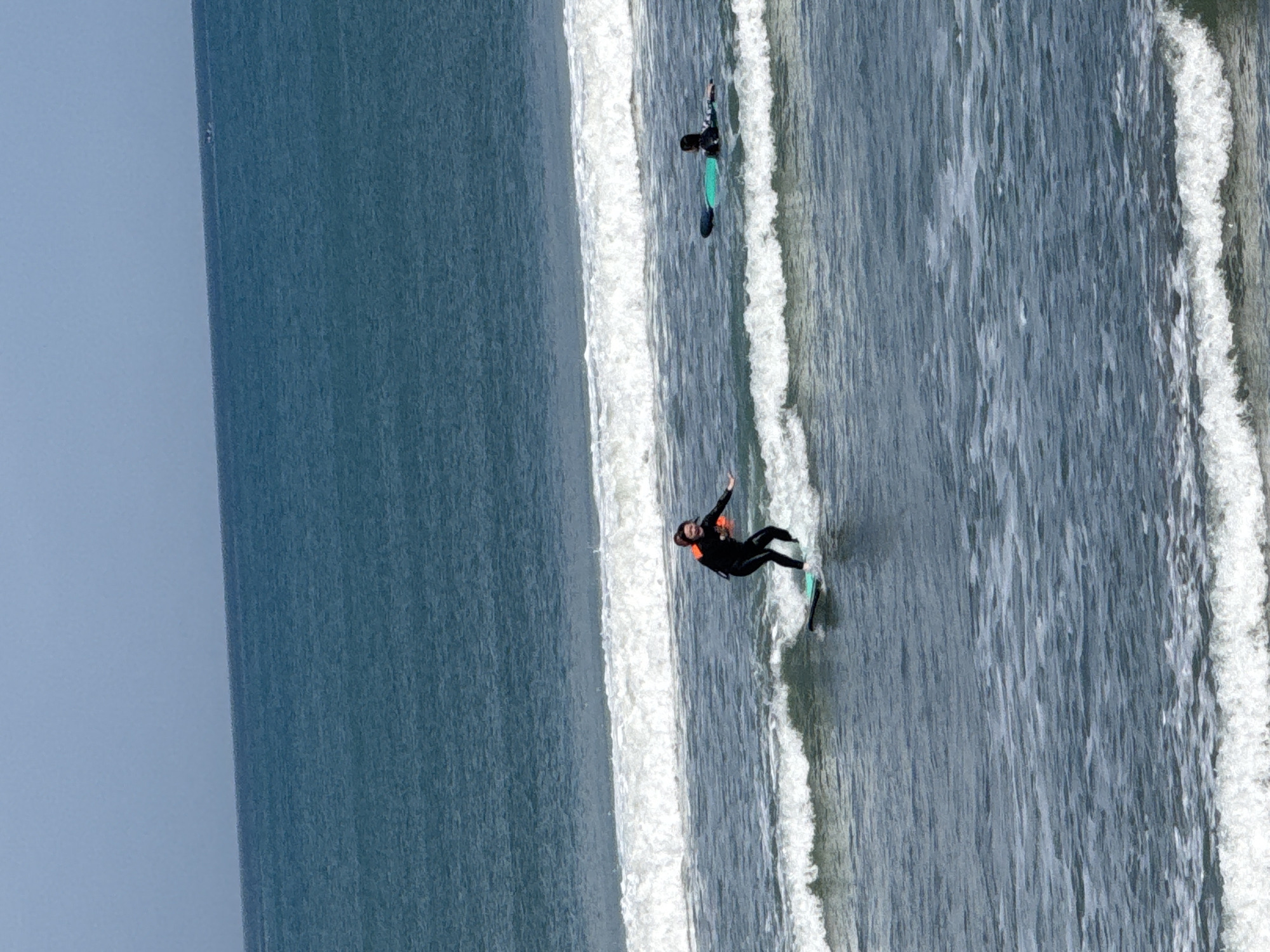 The image depicts two individuals surfing in the ocean. The foreground shows a surfer in a black wetsuit with an orange flotation device on their back, actively riding a wave on a light green surfboard. Behind this surfer, to the right, another person in a black wetsuit is lying on a turquoise surfboard. The ocean is a deep blue with white-capped waves. The horizon is visible in the distance, meeting a light blue sky. There are no visible buildings or landmarks to indicate a specific location within Kuta, Indonesia.