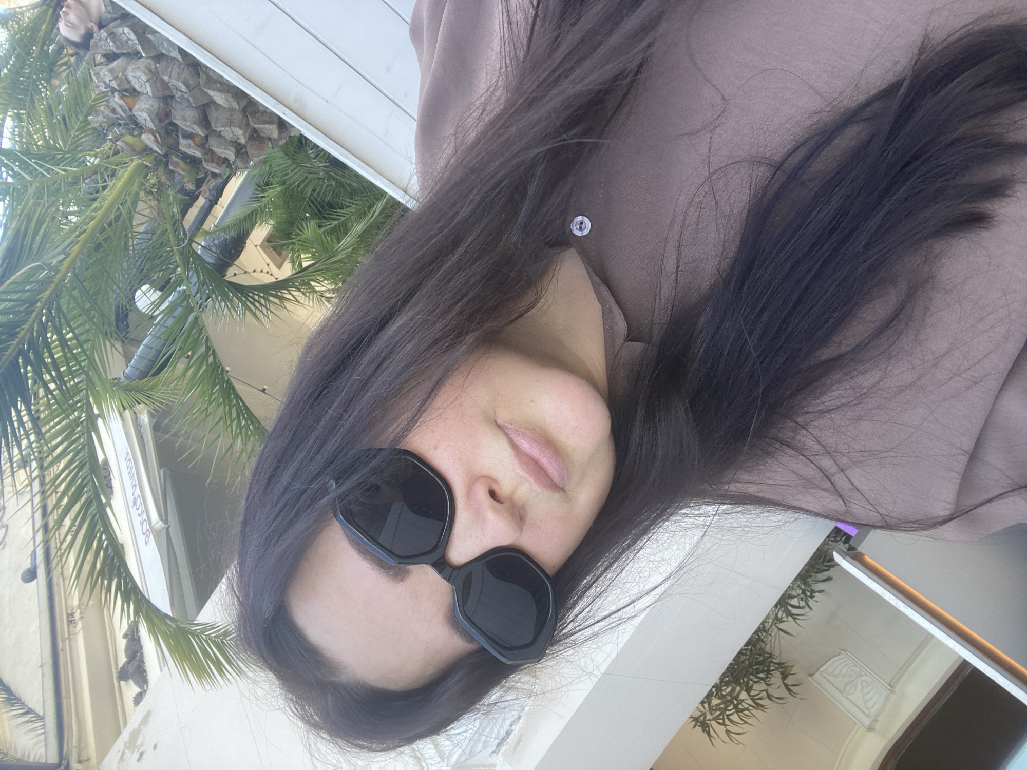 The image features a woman posing outdoors on a sunny day. She is shown from the chest up, with long, dark hair flowing over her right shoulder and partially covering her face. She wears black, angular sunglasses and light pink or nude lipstick. Her attire includes a light brown or taupe-colored collared shirt or blouse, with a single button visible near her collarbone.
In the background, lush green palm trees are visible on the right side of the frame, indicating a warm climate. To the left, a light-colored building with some architectural detailing is present. A white sign affixed to this building clearly displays the text "BOCC RIVIERA". The scene suggests a relaxed, possibly resort-like or urban outdoor setting under bright daylight, consistent with the location context of Sochi, Russia. The woman's expression appears neutral and calm.
