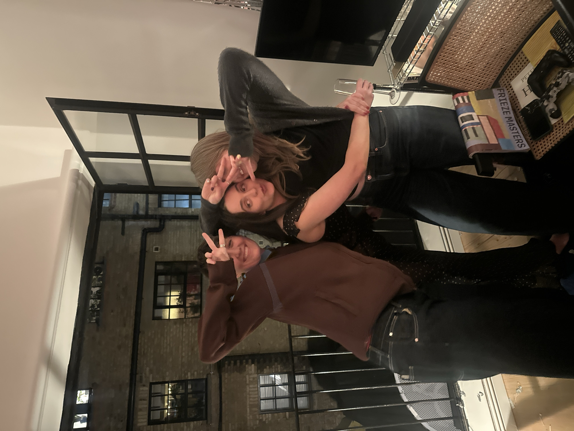 The image shows two people posing indoors at night. One person, wearing a brown sweatshirt, is making a peace sign with their right hand. The other person is making a hand gesture with their hands, framing their face. The person in the brown sweatshirt is standing in front of a building visible through a window, a light-colored brick structure with dark window frames. Inside, there is a television, a glass, and a shelf with books and other objects. The books include one titled "Frieze Masters." Based on the architectural style and the Frieze Masters book, the location is likely in London, United Kingdom.