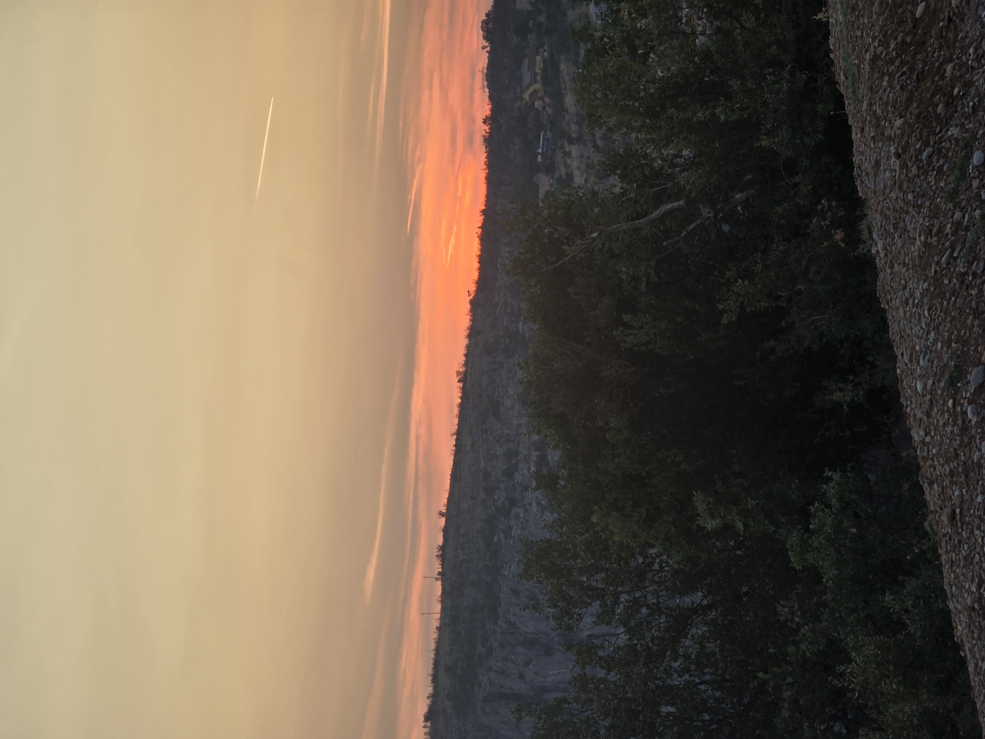 The image depicts a sunset over a hilly, vegetated landscape. In the foreground, there is a pile of small, gravelly stones. Behind the stones, dense dark green foliage forms a barrier across the bottom third of the frame, with some lighter-colored branches intermingled. The middle ground reveals a rocky, sparsely vegetated hillside, with a few trees visible. On the crest of the hill, there are structures that appear to be crosses on poles, and further in the distance, several buildings with visible roofs. The sky is a gradient of warm colors, transitioning from a pale yellow at the top to a vibrant orange and pink near the horizon, with wispy clouds. A faint contrail from an airplane stretches across the upper right portion of the sky. The scene suggests a rural or semi-rural setting in Orljevo, Montenegro, during the evening.