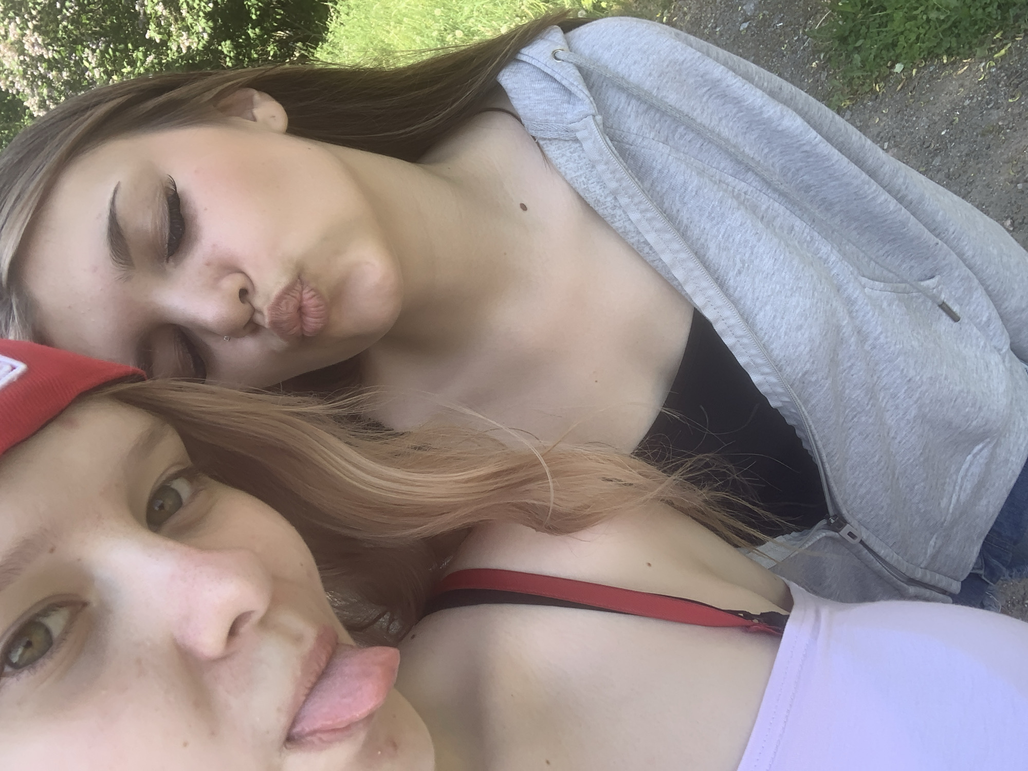 Two young women are captured in a close-up, upward-angled outdoor photograph in Priozersk, Russia. The woman on the left, with fair skin, green eyes, and light brown hair partially covered by a red headwear, extends her tongue playfully towards the camera. She wears a light pink or white top, with a red strap visible on her left shoulder. The woman on the right, positioned slightly behind and to the right, has long brown hair, a small silver nose piercing, and a small brown mole on her right collarbone. She pouts her lips in a 'kissing' expression, with her eyes mostly closed, and wears a black top under a light gray hooded zipper jacket. Blue denim fabric is visible at the bottom edge of the frame. The background features lush green foliage, including trees and bushes adorned with small white or light-colored flowers, and a dirt or gravel path interspersed with patches of grass. The scene is illuminated by natural daylight.