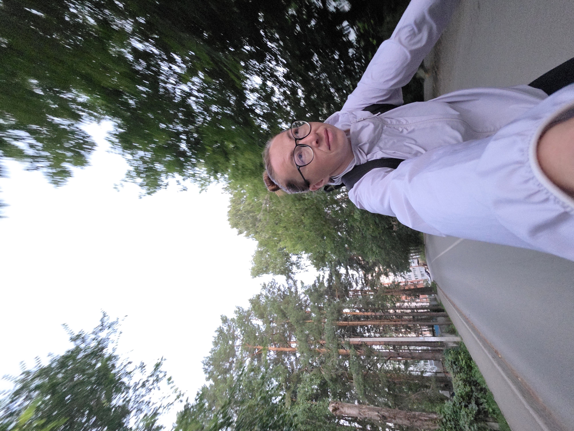 A first-person perspective selfie video taken from a moving vehicle, possibly a bicycle or motorcycle, shows a woman with blonde hair pulled into a bun and wearing glasses and a light-colored jacket. She is looking to her right with a neutral expression. The background reveals a paved road lined with tall trees, indicating a location with a significant amount of greenery. The road appears to be on the outskirts of a city, with some buildings visible in the distance to the left. The overall impression is of a scenic drive through a park-like environment within an urban setting. The timestamp on the video suggests it was captured on August 6, 2023, at 7:55 PM.