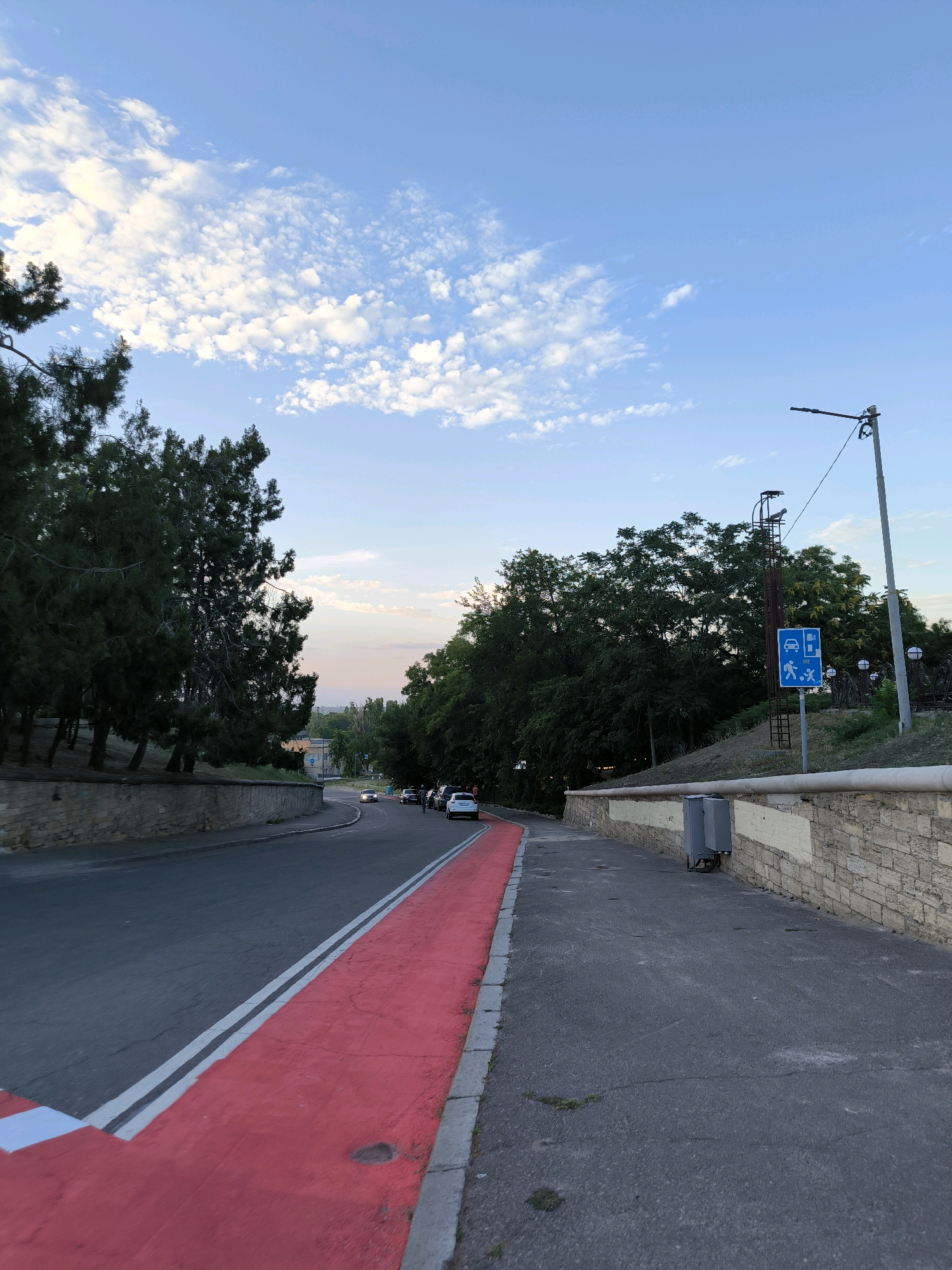 This is a vertical, eye-level, medium shot of a paved road with a bright red lane for bicycles or pedestrians. The road curves to the left and into the distance, where several cars are visible. On the right side of the road, there is a sidewalk and a stone wall, topped with greenery and a signpost. The signpost displays symbols for parking, pedestrians, and possibly a charging station. The scene is set against a backdrop of trees and a pale blue sky with scattered white clouds. The lighting suggests it is late afternoon or early evening. Based on the signage and road layout, this is likely a section of the embankment or a park area in Mykolaiv, Ukraine, possibly near the Inhul River.