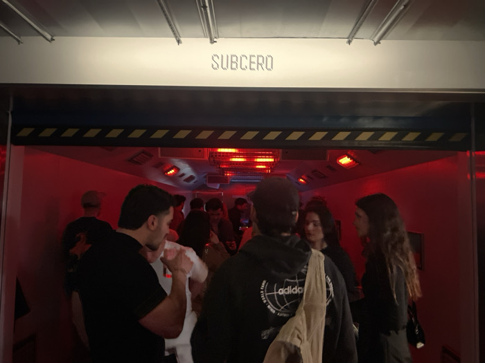 A group of people gather in a room lit with red light under a sign that reads "SUBCERO". Overhead, several heating lamps cast a glow. The room is accessed through a door adorned with a blue and yellow hazard pattern. The group consists of about ten people, many wearing dark clothing, with some conversing or drinking.