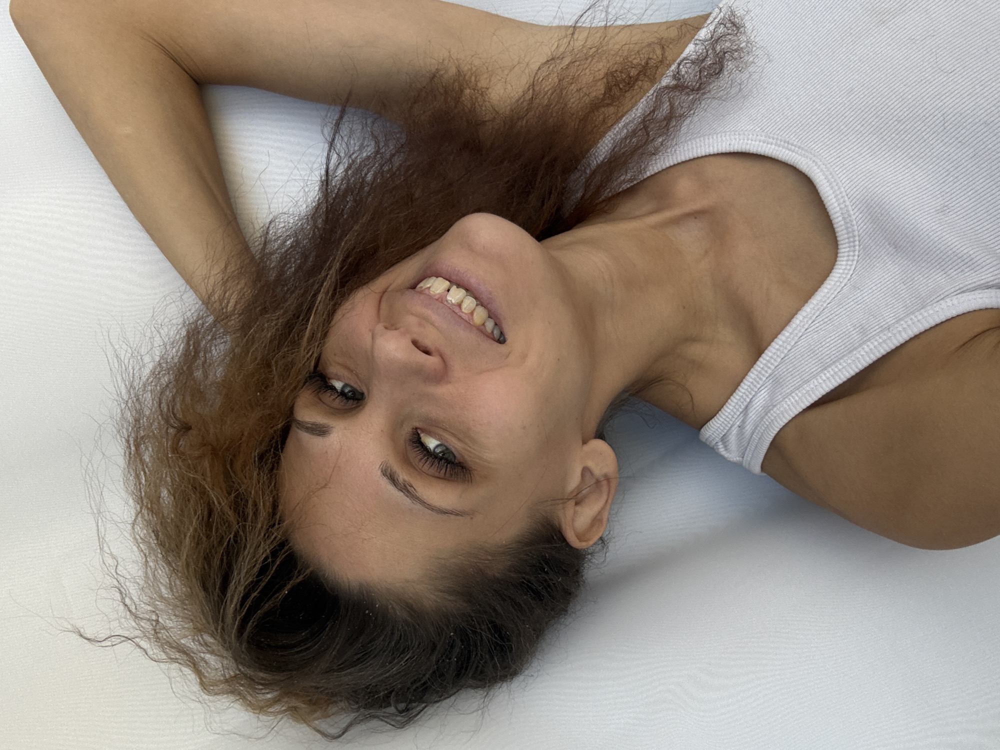 A fair-skinned woman is seen from an overhead perspective. She is lying on a white surface with her head tilted towards the right. She has brown, curly hair that is spread around her head. She is wearing a white tank top. Her arm is raised above her head. She is smiling and looking towards the camera. The scene appears to be indoors. No information indicates that this photo was taken in Rusanovo, Russia.