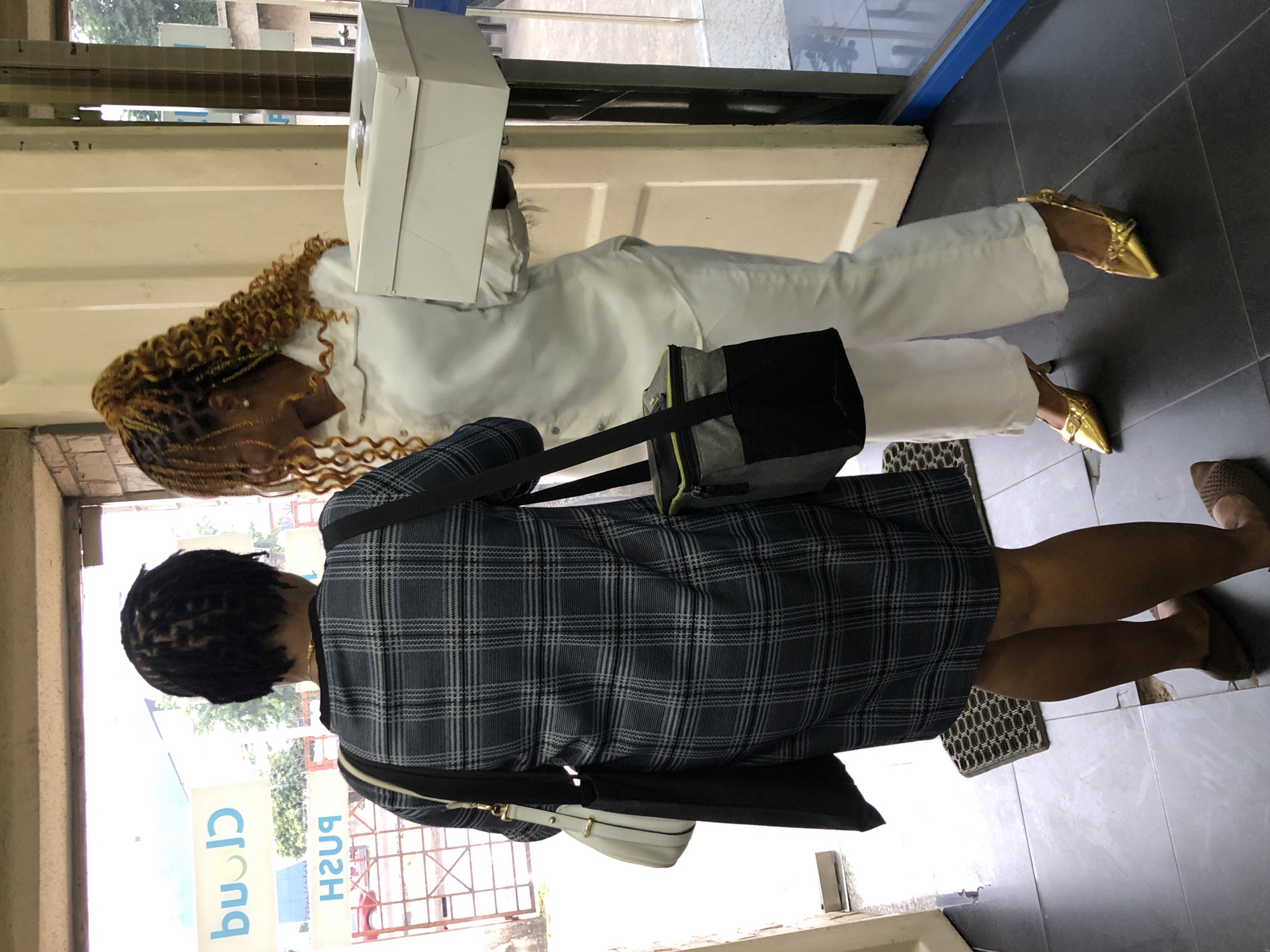 In Ikeja, Nigeria, two women are standing near an open doorway. The woman on the left wears a grey and black plaid dress and a white handbag with a black strap. She has dark dreadlocks and is facing away from the camera. The woman on the right, facing the other woman, wears a white long-sleeved shirt and white pants. Her hair is styled in blonde braids. She is holding a white cake box. The environment appears to be an indoor space with tiled flooring and a mat near the doorway. A sign with the word "Cloud" is visible on a glass panel in the background.