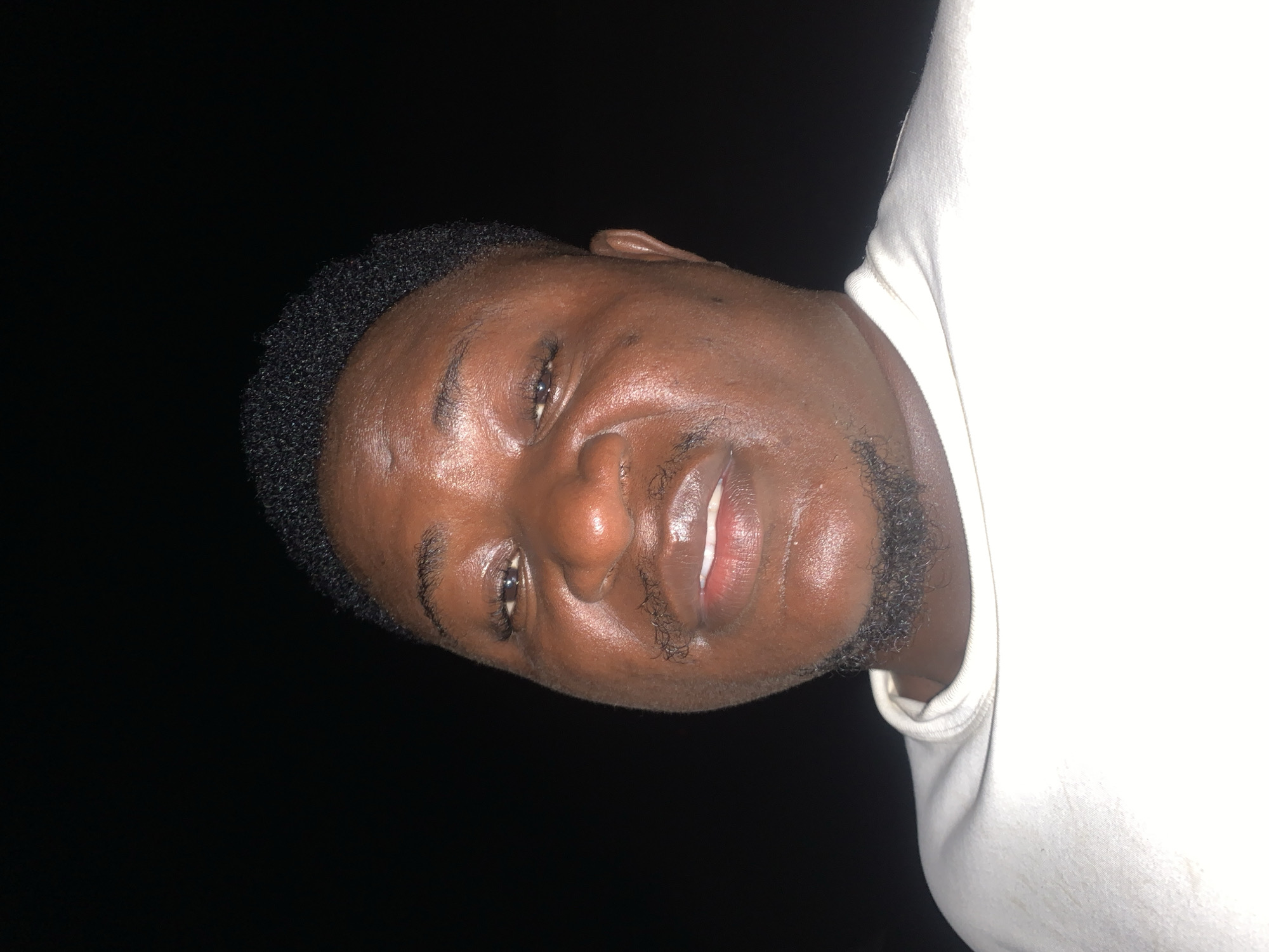 A medium shot taken from a low angle shows a Black man facing the camera. He has dark skin, a round face, and a short, textured hairstyle. He is wearing a white t-shirt. His head is tilted slightly upwards and to his left. He has a goatee and a mustache. The background is black, suggesting an indoor setting or nighttime outdoor setting. No specific landmarks in Akwanga, Nigeria are visible.