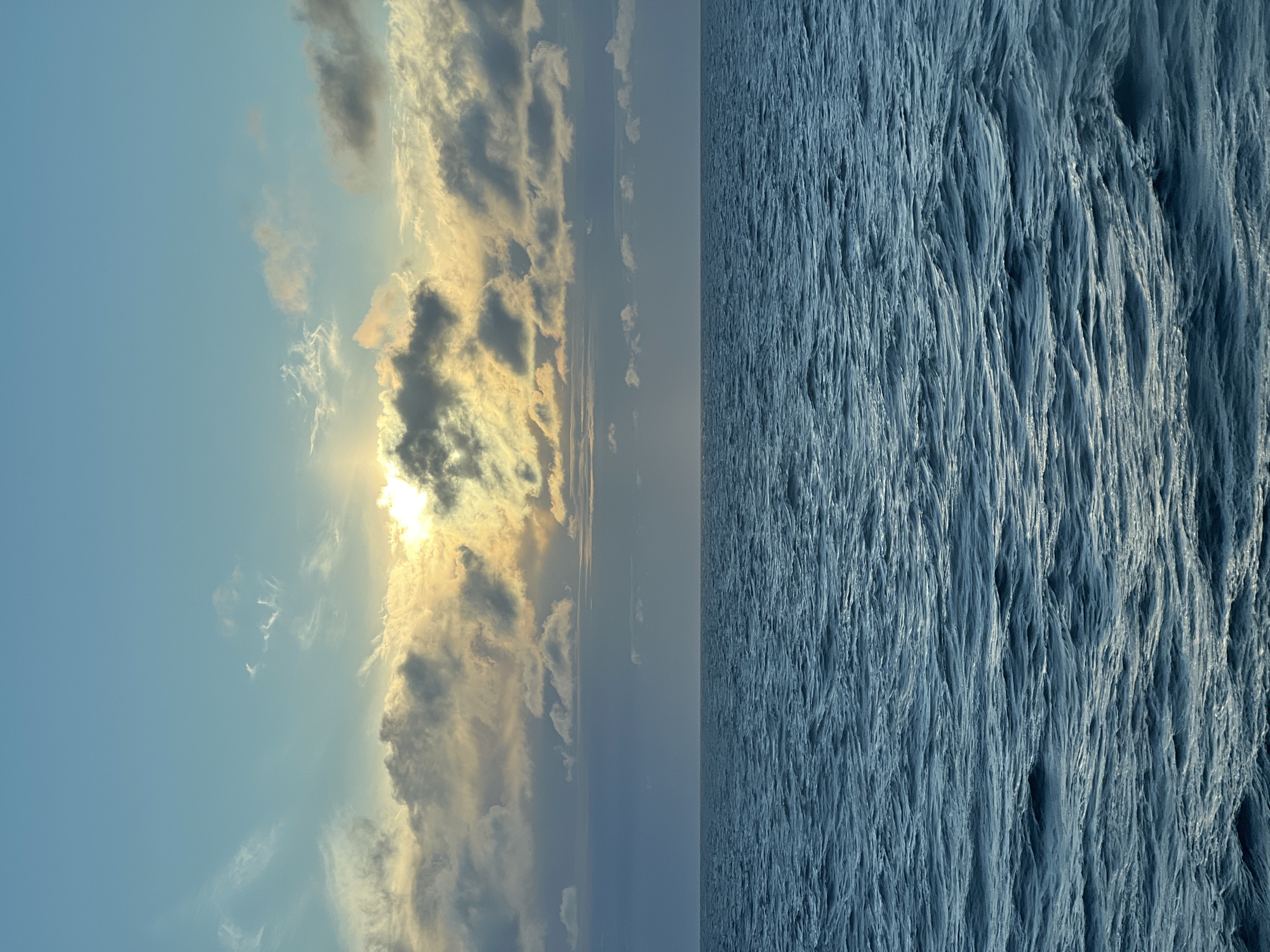 The image captures a serene seascape at sunset. The sky is a gradient of pale blue at the top, transitioning to a soft yellow and orange where the sun is partially obscured by clouds. Wispy cirrus clouds are scattered across the upper portion of the sky, while darker, more textured clouds gather near the horizon. Below the sky lies a vast expanse of deep blue ocean, its surface gently rippling with waves. The horizon line is sharp and clear, dividing the sky from the sea. There are no people, animals, or man-made objects visible in the frame. The scene appears to be an open body of water, possibly the Baltic Sea, given the description of the location being "Unknown, Poland," though the image itself offers no direct clues to confirm this. The overall atmosphere is tranquil and expansive.
