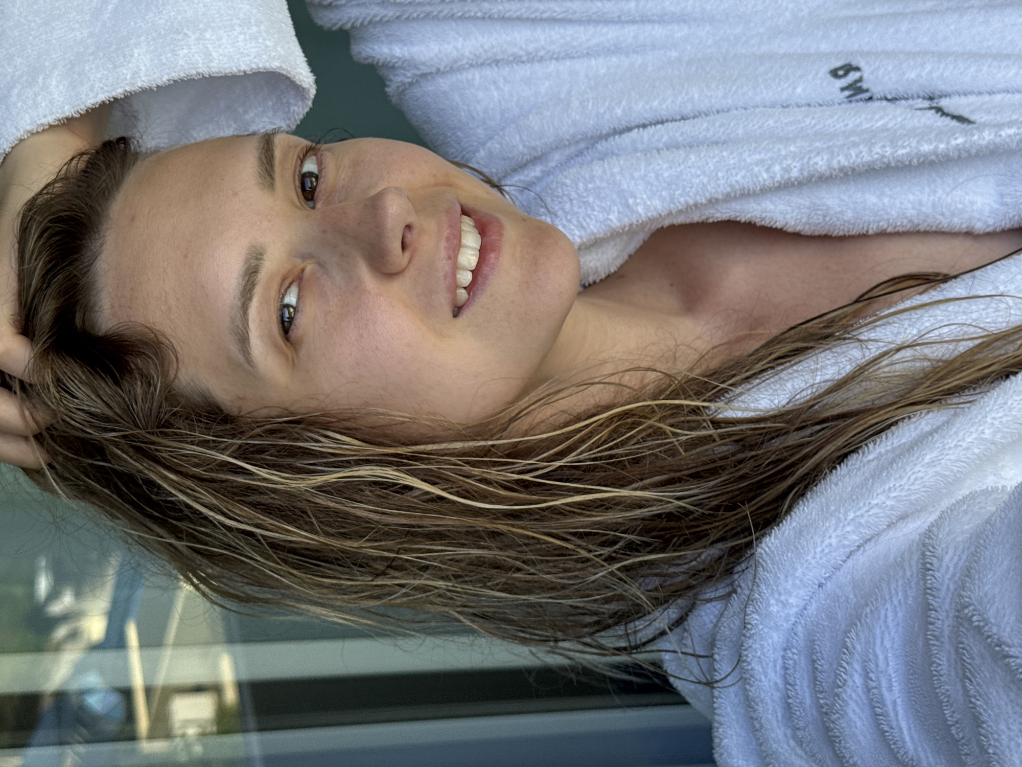 A woman with wet, shoulder-length brown hair and light skin is captured in a close-up, right-facing selfie. She is wearing a white terrycloth bathrobe, and her left hand is raised, touching her hair. The background suggests an indoor setting with a large window reflecting greenery and architectural elements, potentially indicating a hotel or residential building in Nebug, Russia. The lighting is natural and bright, casting subtle shadows on her face.