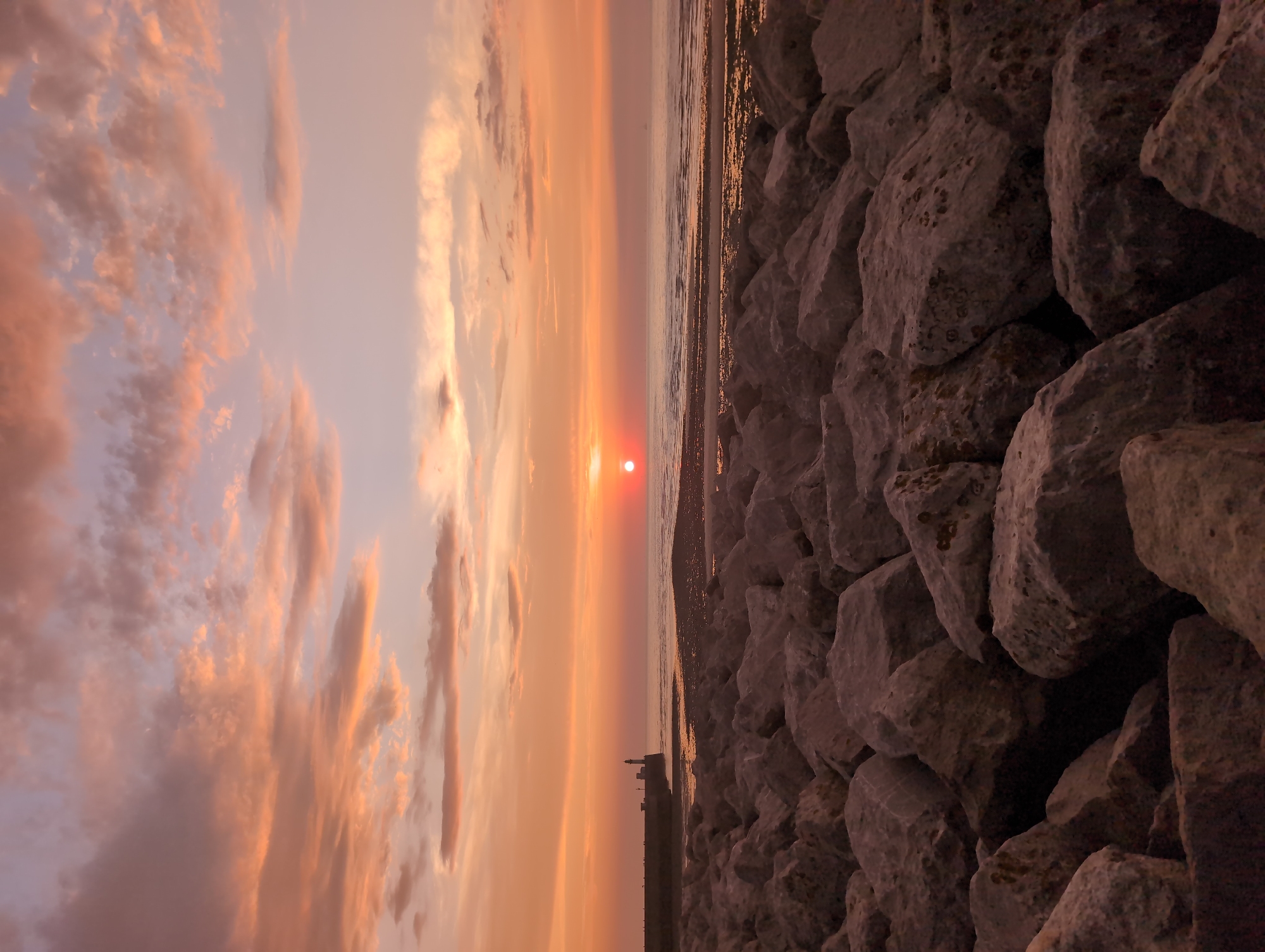 An outdoor, low-angle, vertical, wide shot captures a sunset over the sea at Le Tréport, France. The foreground is dominated by a dense field of large, rough, grey rocks that form a breakwater. The rocks are varied in size and shape, creating a textured and uneven surface.
The middle ground features a calm sea with gentle waves lapping at the shore. The wet sand reflects the warm colors of the sunset. A long, dark pier stretches out into the water from the left side of the frame. At the end of the pier stands a tall, slender lighthouse, its white structure stark against the colored sky.
The background is a dramatic sunset sky filled with clouds. The clouds are tinged with shades of orange, pink, and lavender, with the sun a small, bright orange orb just above the horizon. The colors suggest the final moments of daylight. There are no people or animals visible in the scene.