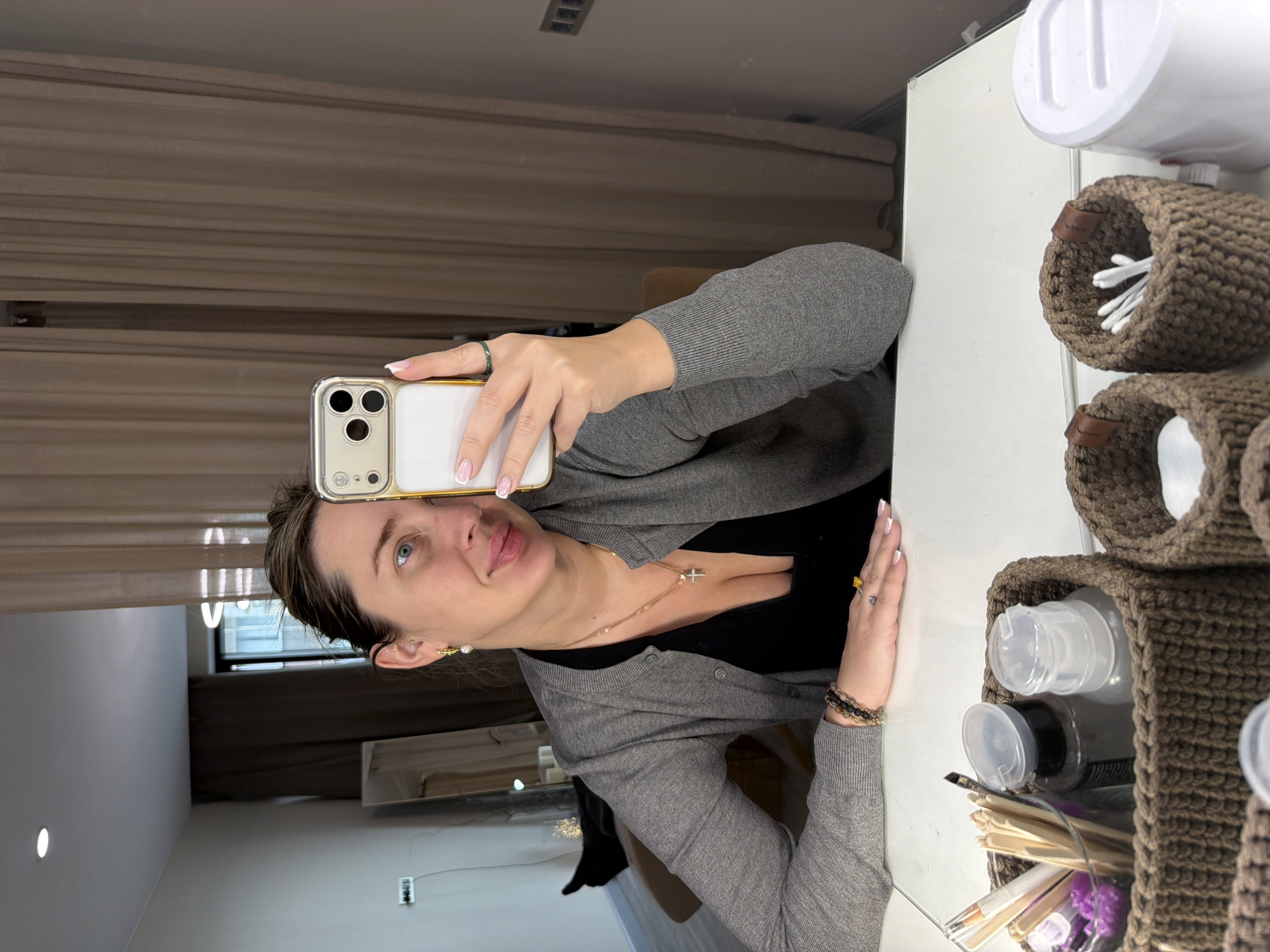 A woman is taking a selfie in a mirror. She is holding a gold iPhone with a white case. She is wearing a gray cardigan over a black shirt. She has brown hair pulled back, and is wearing a gold necklace with a cross pendant. Her fingernails are light pink. The background is a room with curtains and a window. On the right side of the image, there are several brown crocheted containers holding items such as cotton swabs and bottles. There is no evidence to indicate that the photo was taken in Khimki, Russia.