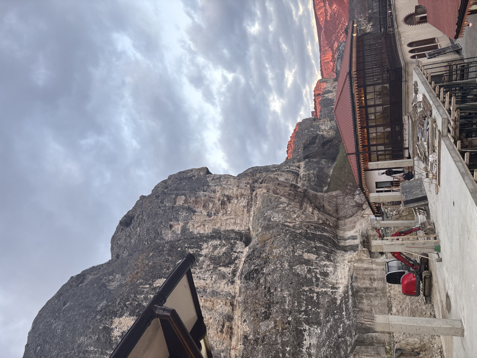 The image displays a construction site at the Holy Dormition Cave Monastery (Uspensky Monastery) in Bakhchysarai, Ukraine, situated directly against a prominent, weathered grey and tan limestone cliff. The sky is uniformly overcast with grey clouds. In the mid-ground, a concrete slab is under construction, supported by wooden scaffolding and several unfinished concrete pillars. A compact red excavator with a grey cab and bucket arm is parked on disturbed earth at the base of the cliff, positioned behind the concrete work. To the right, an existing two-story building features a red gabled roof, light-colored walls, and an enclosed upper-level balcony with dark wooden latticework and multi-paned windows. Adjacent to this structure, a simpler, single-story light-colored building has two individuals dressed in dark attire standing near its entrance. In the background, distant cliffs and mountains are bathed in a distinct reddish-orange light, indicating a low sun angle. The scene depicts ongoing construction or renovation activity integrated into a historical mountainous landscape.