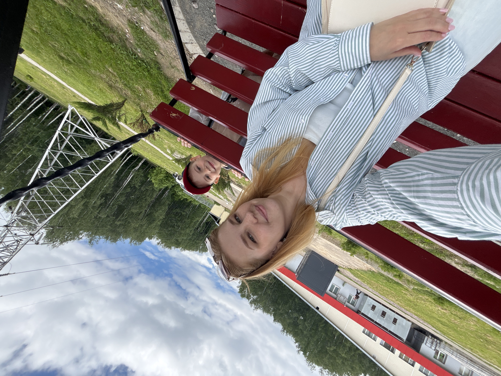 The image portrays an outdoor scene featuring two individuals, a young woman and a child, seated on a red slatted bench. The primary subject, a young woman with blonde hair, occupies the foreground, looking directly at the camera with a gentle smile, suggesting she is taking a selfie. She is dressed in a light blue and white striped long-sleeved shirt worn over a white top, and a light-colored shoulder bag is visible on her lap.
Behind her, partially obscured by the woman, a child wearing a red baseball cap is also visible, looking towards the camera with a slight smile. Both appear relaxed, enjoying the outdoor setting.
The bench is positioned alongside a gravel or dirt path. Beyond this, a wide expanse of green grass extends towards a backdrop of lush deciduous trees, likely a forest. A prominent metal lattice tower, possibly part of an electrical transmission line, stands on the left side of the frame, with power lines stretching across the sky.
The sky above is partly cloudy, featuring large white clouds against patches of blue. In the middle ground, on the right side, several buildings are visible, including one with a distinctive red roofline and another multi-story grey structure with numerous windows. The overall impression is a daytime scene with pleasant weather, likely late morning or afternoon. The location cues, including the natural landscape, infrastructure, and simple buildings, are consistent with a semi-rural or rural setting, such as Podosenikha, Russia. There is no visible text in the image.