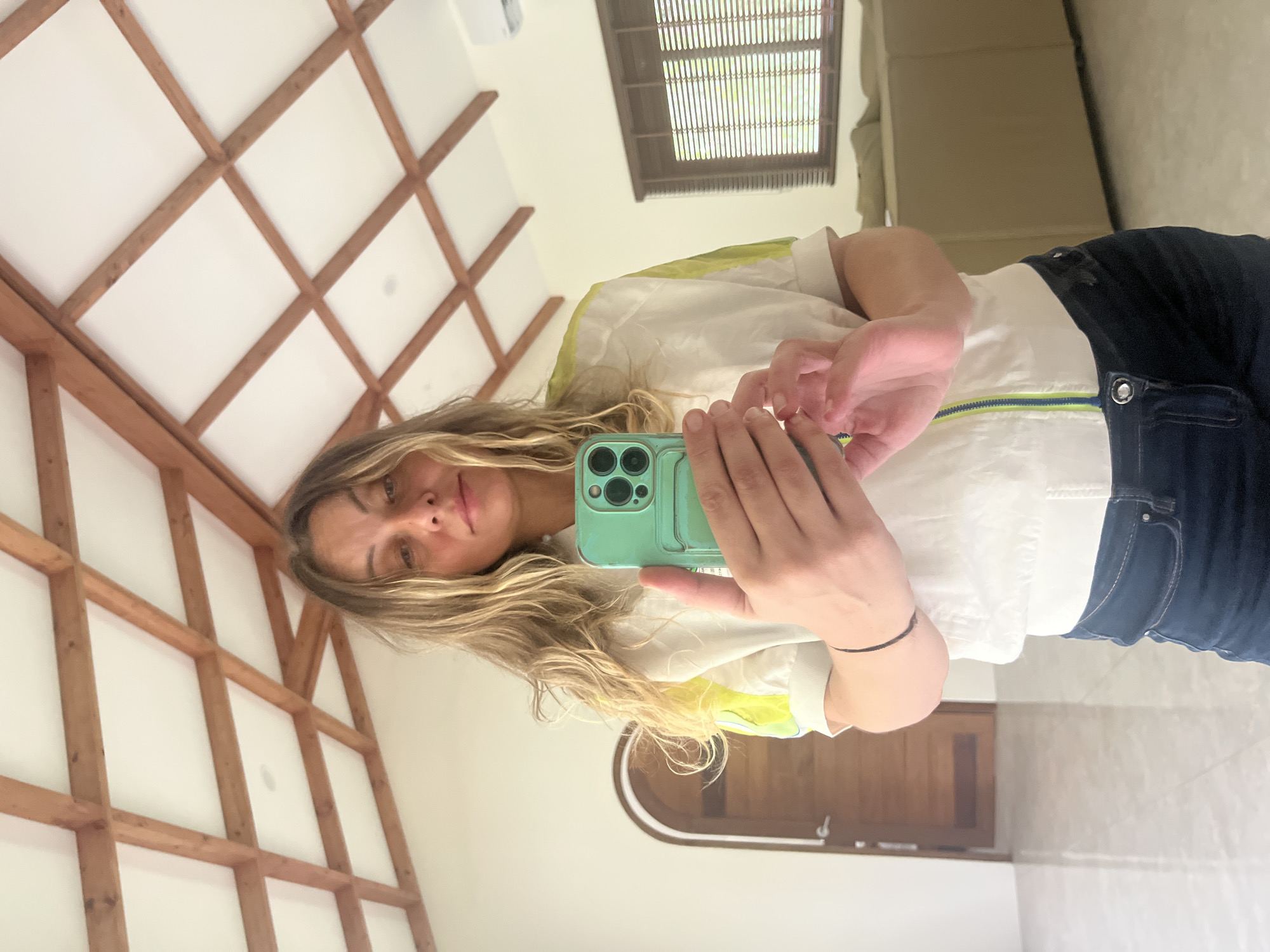 A woman takes a mirror selfie in a room with a white ceiling featuring exposed wooden beams. She is wearing a white jacket with neon green accents and dark blue jeans. Her hair is blonde and wavy. In her right hand, she holds a mint green smartphone, capturing her reflection. To the left of the frame, a tall wooden door with an arched top is visible. To the right, a window with horizontal blinds is present, and in the background, a light brown sofa can be seen. The floor is a light-colored, reflective tile. The overall ambiance suggests a residential interior in a tropical location, consistent with the description of Koh Phangan, Thailand.
