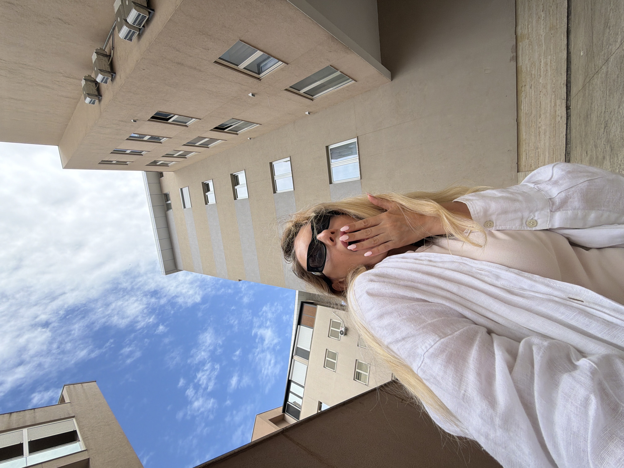 A low-angle, upward-facing shot in an urban environment, possibly in Budva, Montenegro, features a fair-skinned woman with blonde hair wearing sunglasses and a white linen shirt. She is positioned to the right, and part of her face is covered by her hand. The architectural environment includes multiple buildings with light-colored facades and several windows. The sky, featuring a clear blue color with some clouds, is visible between buildings. The presence of air conditioning units on one of the buildings is noticeable.