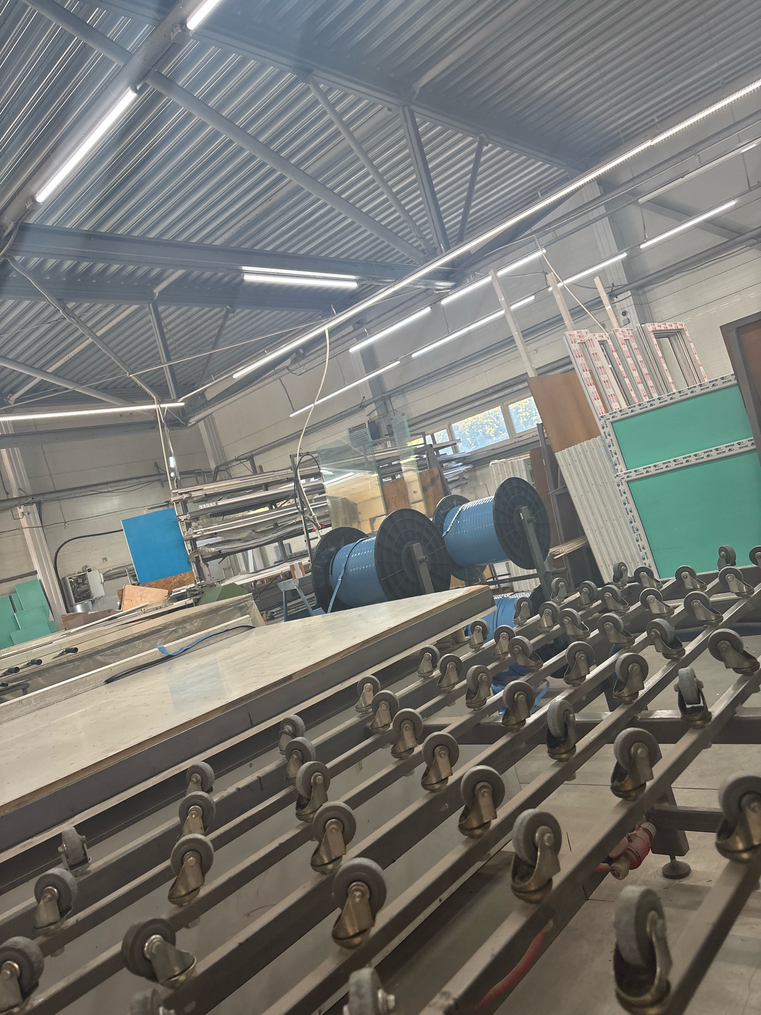 The image depicts an interior view of a workshop or factory setting. The ceiling is constructed of corrugated metal, illuminated by long, white fluorescent or LED lights. Several steel beams support the structure. In the midground, large blue spools are visible, likely used for storing materials like cable or wire. To the right, stacks of what appear to be window frames, possibly made of PVC, are leaning against a wall. The foreground is dominated by a conveyor system with numerous small, wheeled rollers. Some of these rollers are dark grey, while others have a brass-colored swivel base. A red object, possibly a tool or a piece of equipment, is situated on the conveyor in the lower right portion of the image. The overall environment suggests an industrial or manufacturing facility. No people are visible in the image, and no specific location in Barnaul, Russia, can be identified from the visual information provided.
