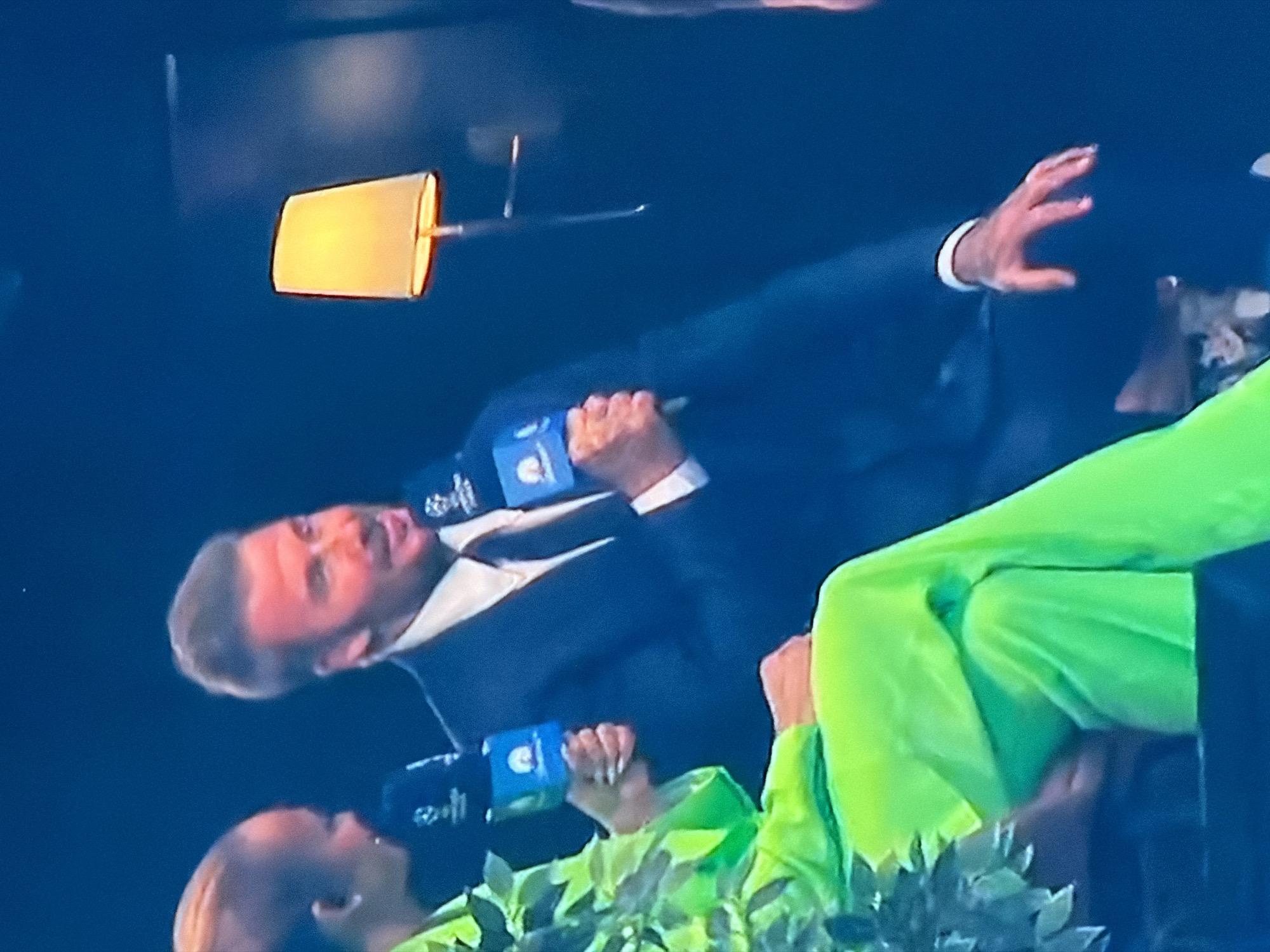 The image shows two individuals holding microphones with the UEFA Champions League logo. One person is wearing a dark suit and white shirt, while the other is wearing bright green clothing. A lamp with a gold shade is visible in the upper part of the image. No evidence indicates the location is Asenovgrad, Bulgaria.