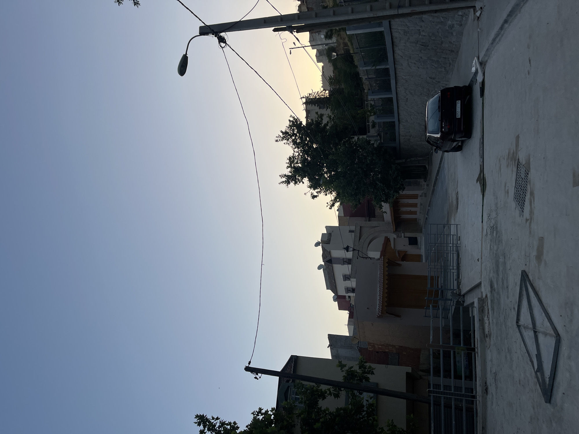 The image depicts a street scene in El Hajeb, Morocco, during twilight. The sky is a clear gradient of pale blue to pale yellow, indicating the setting sun. Buildings, with their characteristic flat roofs and satellite dishes, line the street. Some buildings have arched doorways and wooden shutters. A dark-colored sedan is parked on the right side of the street, facing away from the viewer, with its license plate visible. A street lamp is present on the right, and power lines crisscross the sky. In the foreground, a metal frame lies on the paved ground, and a metal grate is visible in the pavement. Trees with dense foliage are scattered throughout the scene, adding greenery to the urban environment. The overall atmosphere is quiet and serene, typical of a residential area at dusk.