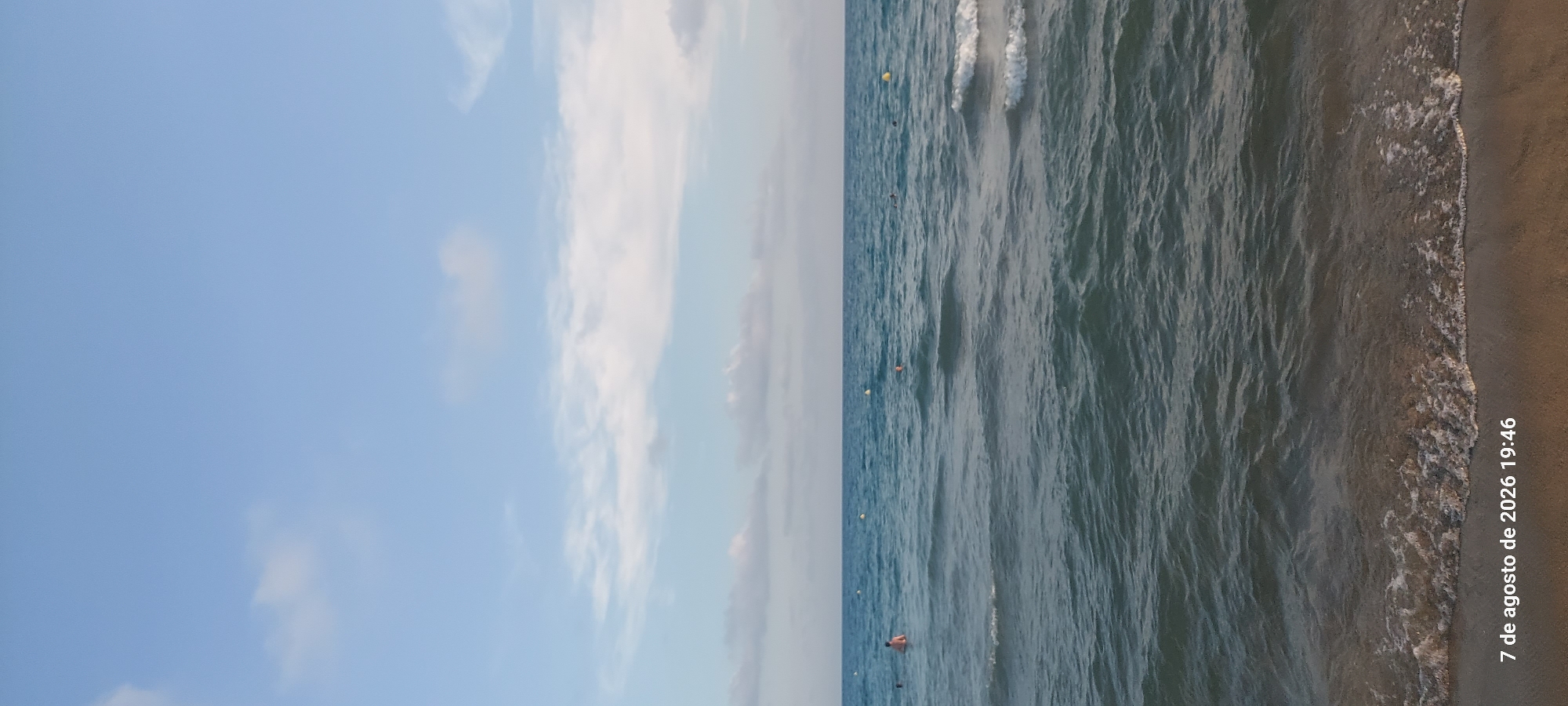The image captures a serene seascape at the beach of Castelldefels, Spain, as indicated by the timestamp "7 de agosto de 2026 19:46." The upper portion of the frame is dominated by a pale blue sky with scattered, wispy white clouds, suggesting it is late afternoon. Below the sky, the calm, deep blue sea stretches to the horizon, with subtle ripples and waves indicating a gentle breeze. Several yellow buoys are visible in the water, marking a designated swimming area. In the mid-ground, a few individuals are present in the sea. One person is partially submerged, their torso visible above the water, and another appears to be swimming further out. The foreground shows the sandy shore meeting the water's edge, with small waves gently lapping onto the beach, creating a foamy white line. The overall atmosphere is peaceful and indicative of a typical summer evening at the coast.
