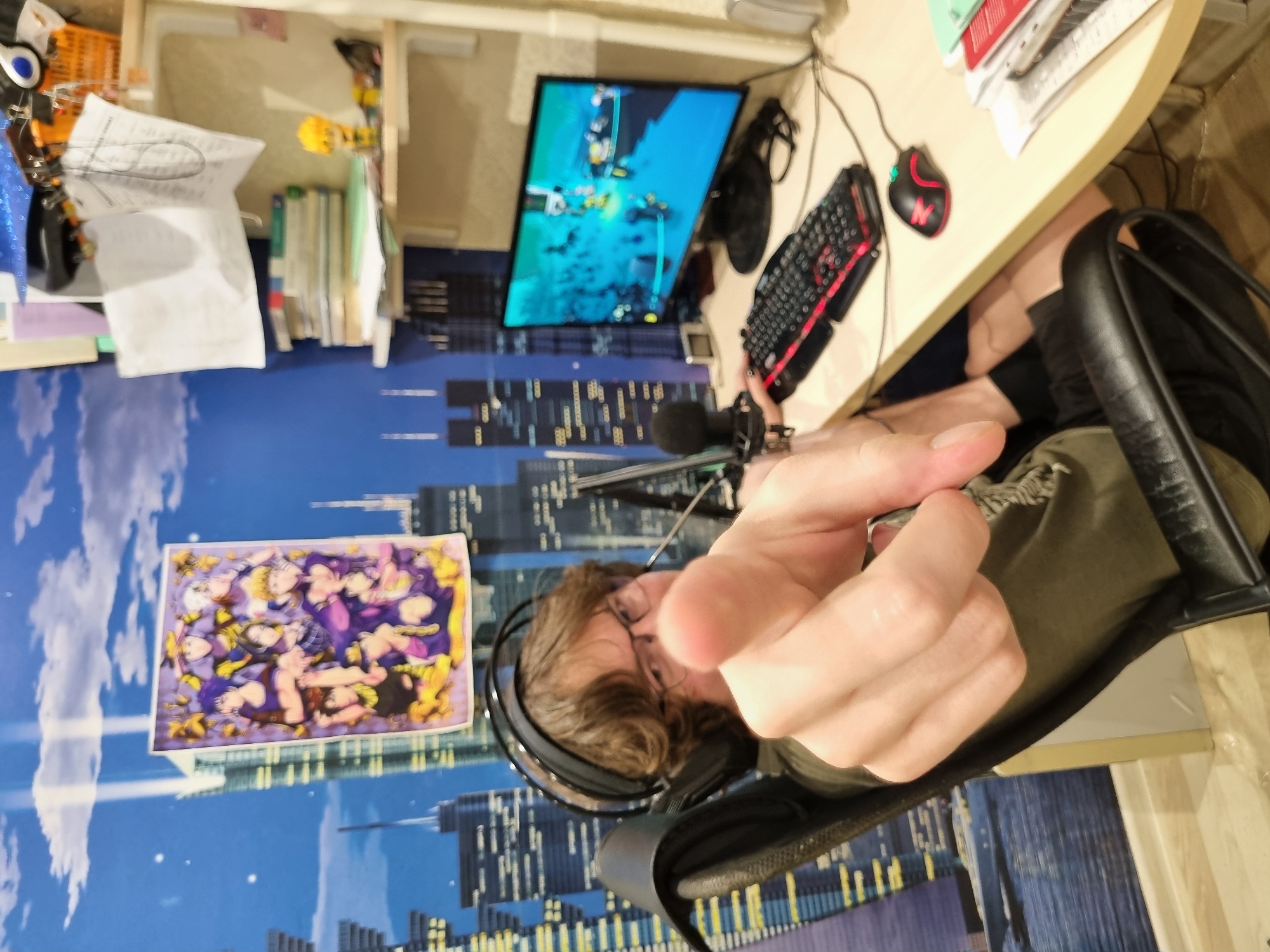 The image shows a person sitting at a desk with computer equipment. The desk has a monitor displaying a video game, a keyboard, a mouse, and a microphone. The person is wearing headphones. A picture is on the wall behind the desk. The background suggests a city skyline with skyscrapers.
The location is not discernable as Chaikovsky, Russia.