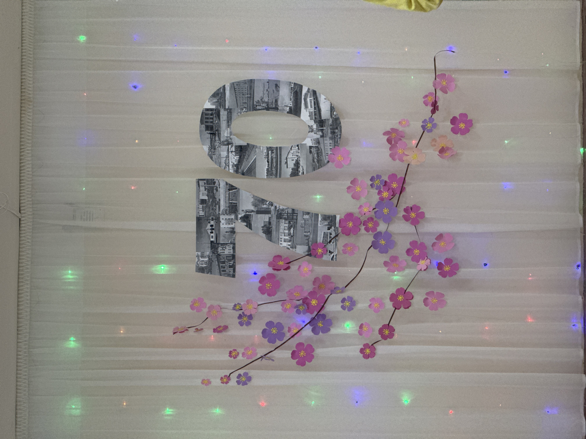 The image depicts a decoration on a white, possibly fabric-covered wall or backdrop. The main feature is a large, stylized "20" formed from black and white photographs of urban landscapes, likely cityscapes with buildings and streets. A decorative branch with pink and purple paper flowers is arranged diagonally across the "20." Small, colorful lights are scattered across the backdrop, adding to the festive or decorative nature of the scene. The environment suggests an indoor setting, possibly a home or event space.