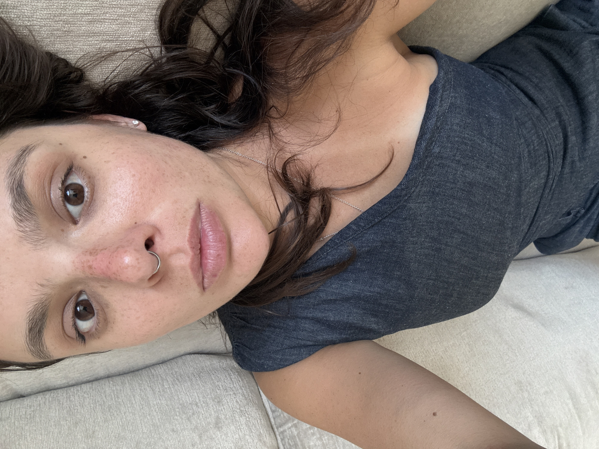 An overhead portrait features a young woman lying on a light-colored, textured fabric couch or bed, looking directly upwards into the camera. Her head is positioned towards the bottom right of the frame, with her body extending diagonally towards the upper left. She has dark brown eyes, slightly parted pink lips, and a silver hoop piercing in her nose. Numerous freckles are visible across her nose and cheeks. Her dark, wavy hair cascades around her head. She wears a dark blue or charcoal grey casual top and a thin silver chain necklace. A small diamond stud earring is visible in her right earlobe. Her skin exhibits a light tan with some faint tan lines on her upper chest and shoulder. A small mole is visible on her upper left arm/shoulder area. The background consists entirely of the same light-colored, woven fabric of the furniture.