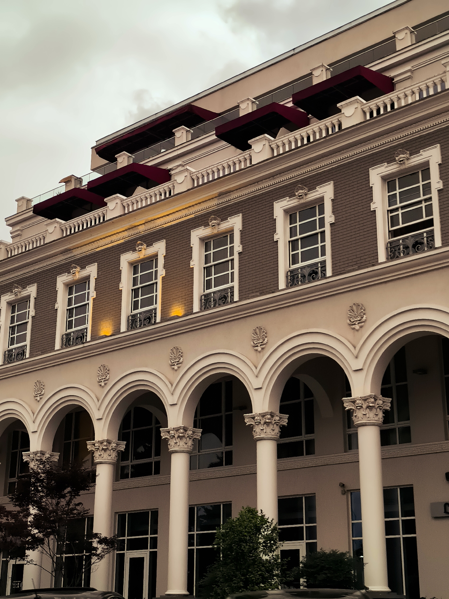 The image captures the facade of a grand building with a classical architectural style, located in Batumi, Georgia. The building features a beige and grey color palette with ornate white trim. The upper floors are characterized by several evenly spaced, tall windows with white frames and decorative ironwork, illuminated from below by warm yellow lights. Above these windows, a row of red awnings is visible on the top floor, suggesting balconies or terraces.
The ground floor is defined by a series of white arches supported by fluted columns topped with Corinthian capitals. Behind these arches, large windows and doorways are visible, some reflecting the cloudy sky. Lush green trees and dark foliage are present in front of the lower level, obscuring some of the ground-floor details. The sky is overcast, contributing to a subdued and atmospheric lighting condition. The overall impression is one of elegance and historical grandeur, typical of upscale urban architecture.