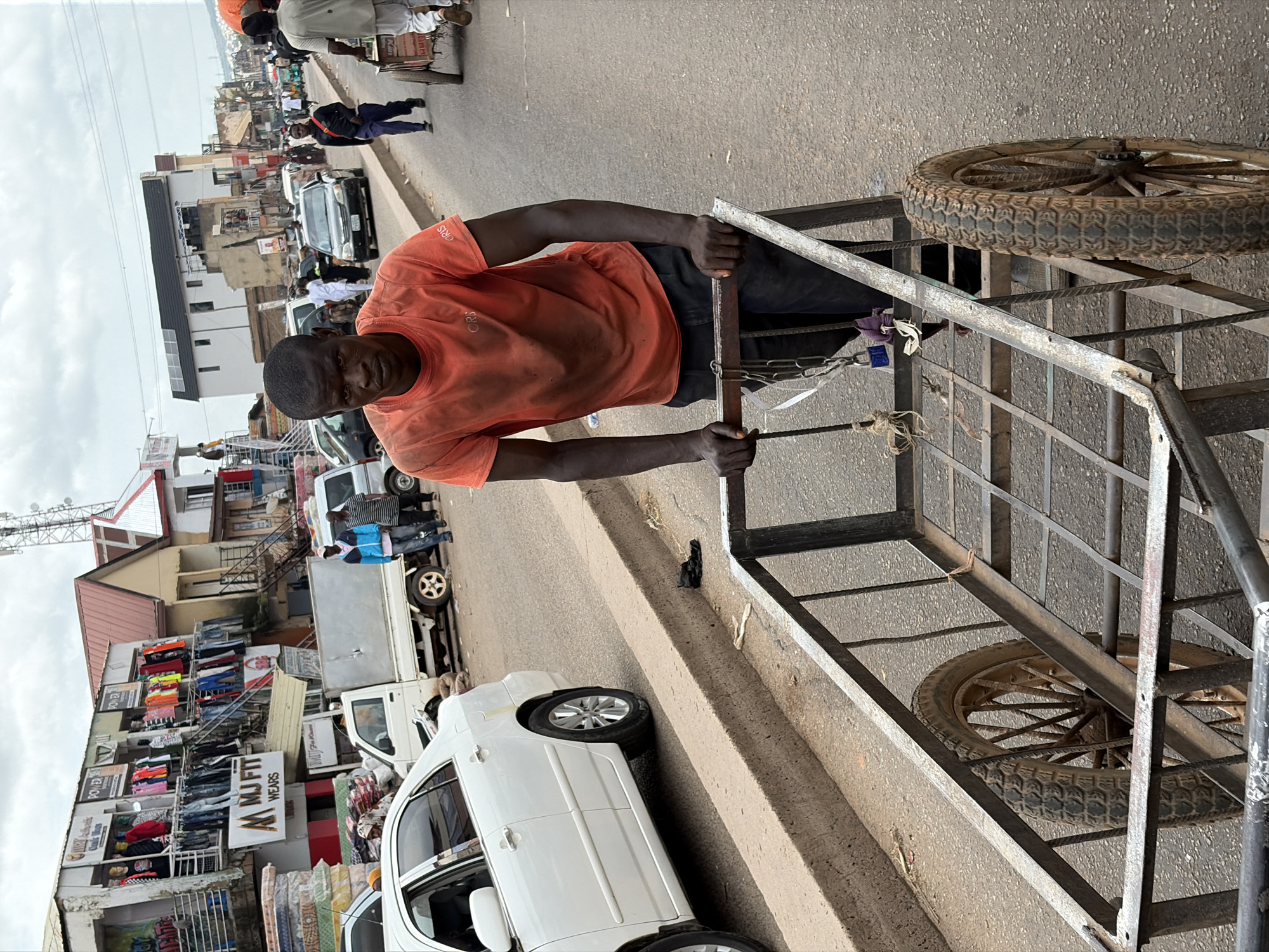 The image depicts a street scene in Jos, Nigeria, under an overcast sky. In the foreground, a Black man wearing an orange t-shirt and dark trousers is pushing a metal utility cart with two large spoked wheels. He is facing forward and looking directly at the camera. The cart is laden with what appears to be sacks and fabric. The man's hands are on the cart's handles, and he is positioned on a paved street.
Behind the man, to the left, a white sedan car is parked at an angle on the side of the road. Further back, a white utility truck with a cargo bed is also visible. The background features a row of buildings, some appearing to be shops, with clothing and other merchandise displayed outside. These buildings suggest a commercial area. Several other people are walking or standing in the middle and background of the scene, some appearing to be customers or vendors. The overall impression is one of a busy street market or commercial district.