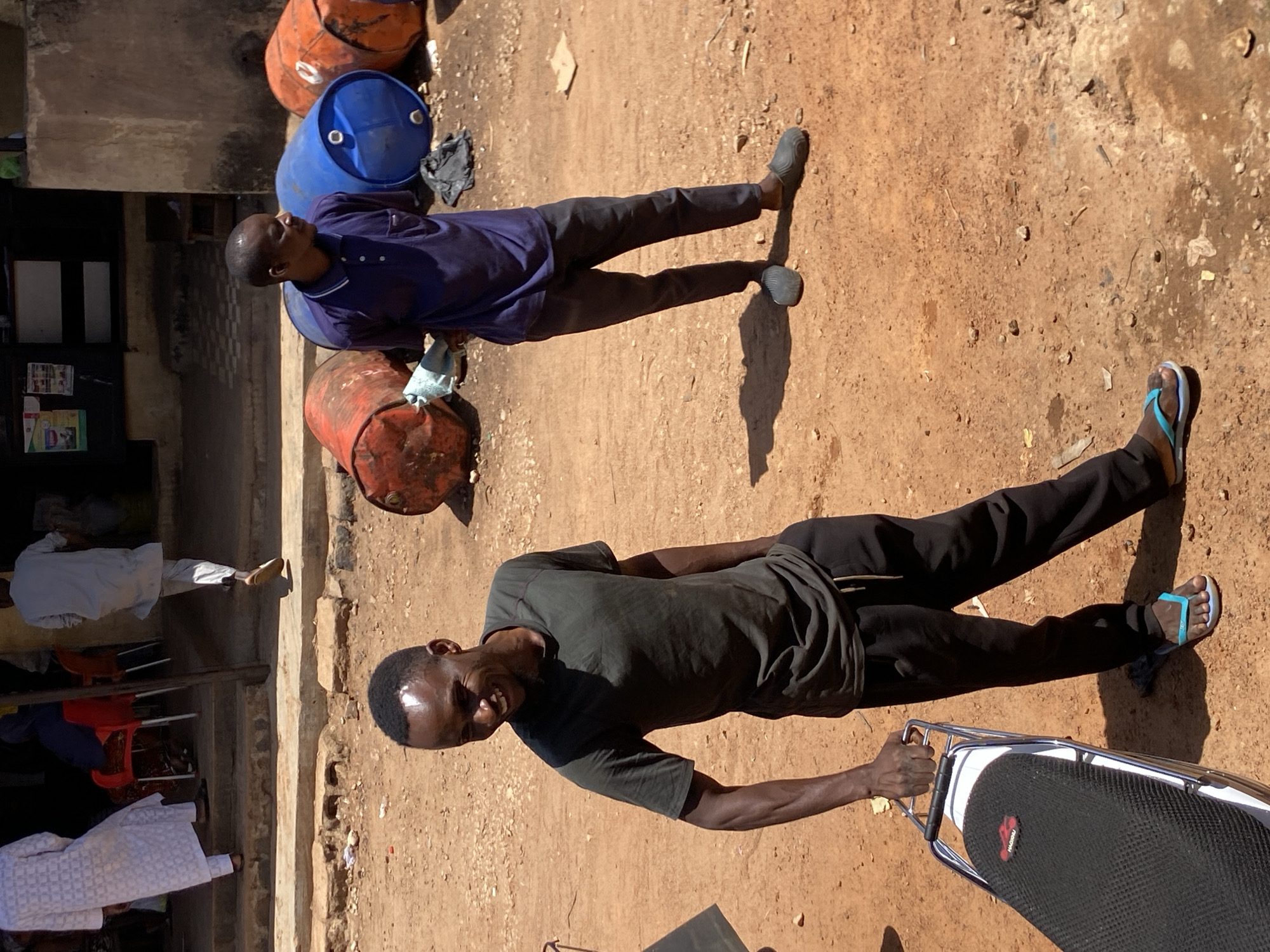 The image depicts an outdoor scene in Bukuru, Nigeria, characterized by a dirt ground with scattered debris. Two men are prominently featured. One man is wearing a dark green t-shirt and black pants, with blue flip-flops, and is holding onto the back rack of a motorcycle. The second man is wearing a purple collared shirt and dark pants, and wearing dark colored slip-on shoes. Several large metal barrels are scattered in the background. The barrels are blue, orange, and rusty red. The building behind the barrels is made of concrete.
