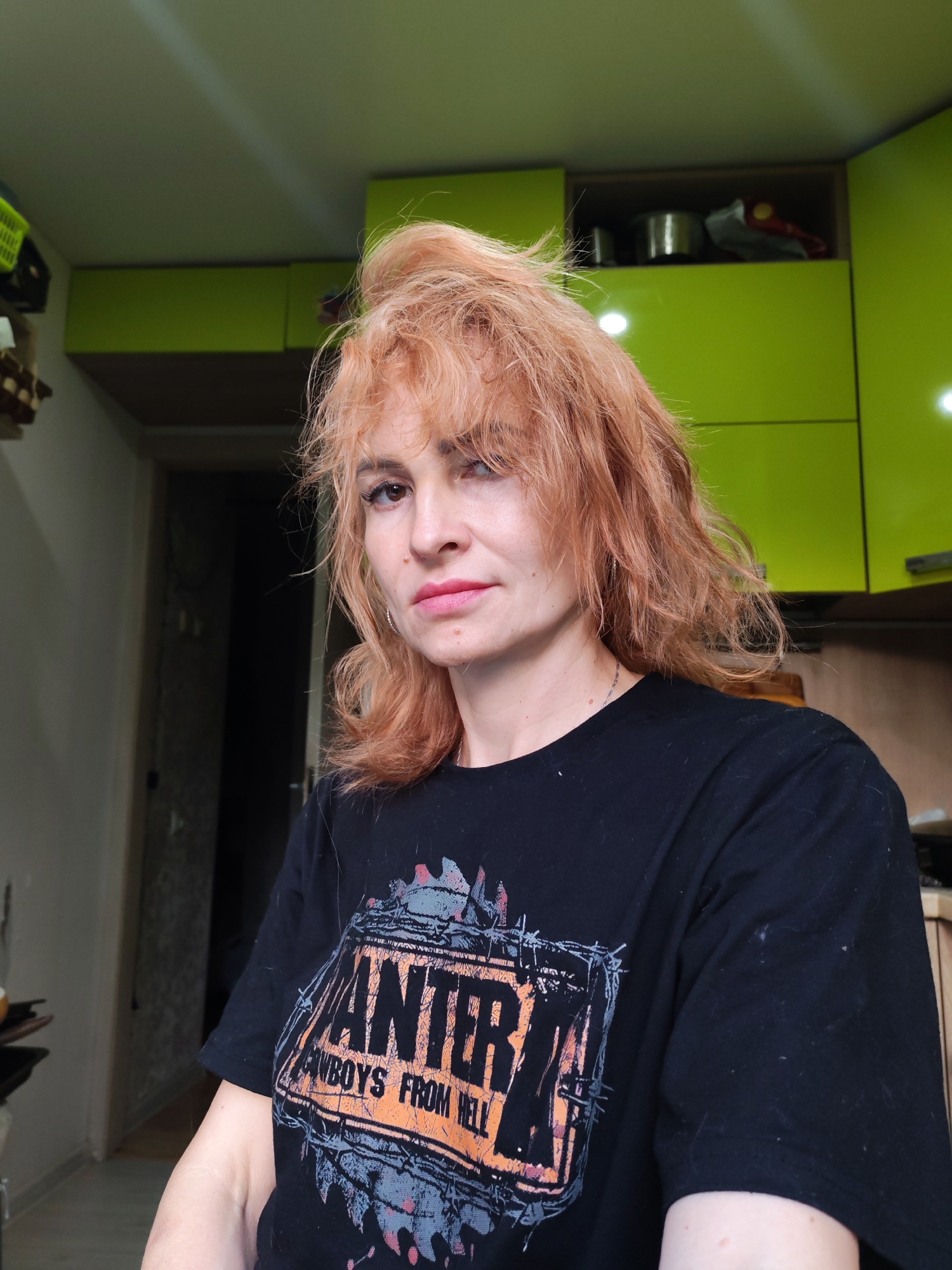 The image shows a woman with reddish-blonde, messy hair and light skin, wearing a black t-shirt with a graphic print. She is looking towards the camera with a neutral expression. The background appears to be an indoor setting, possibly a kitchen, with bright green cabinets. The lighting suggests it is daytime, with a natural light source and possibly some artificial lighting. The overall impression is of a casual, candid photograph. The location of Chaikovsky, Russia, is mentioned, but there are no visual cues in the image that definitively place it there.