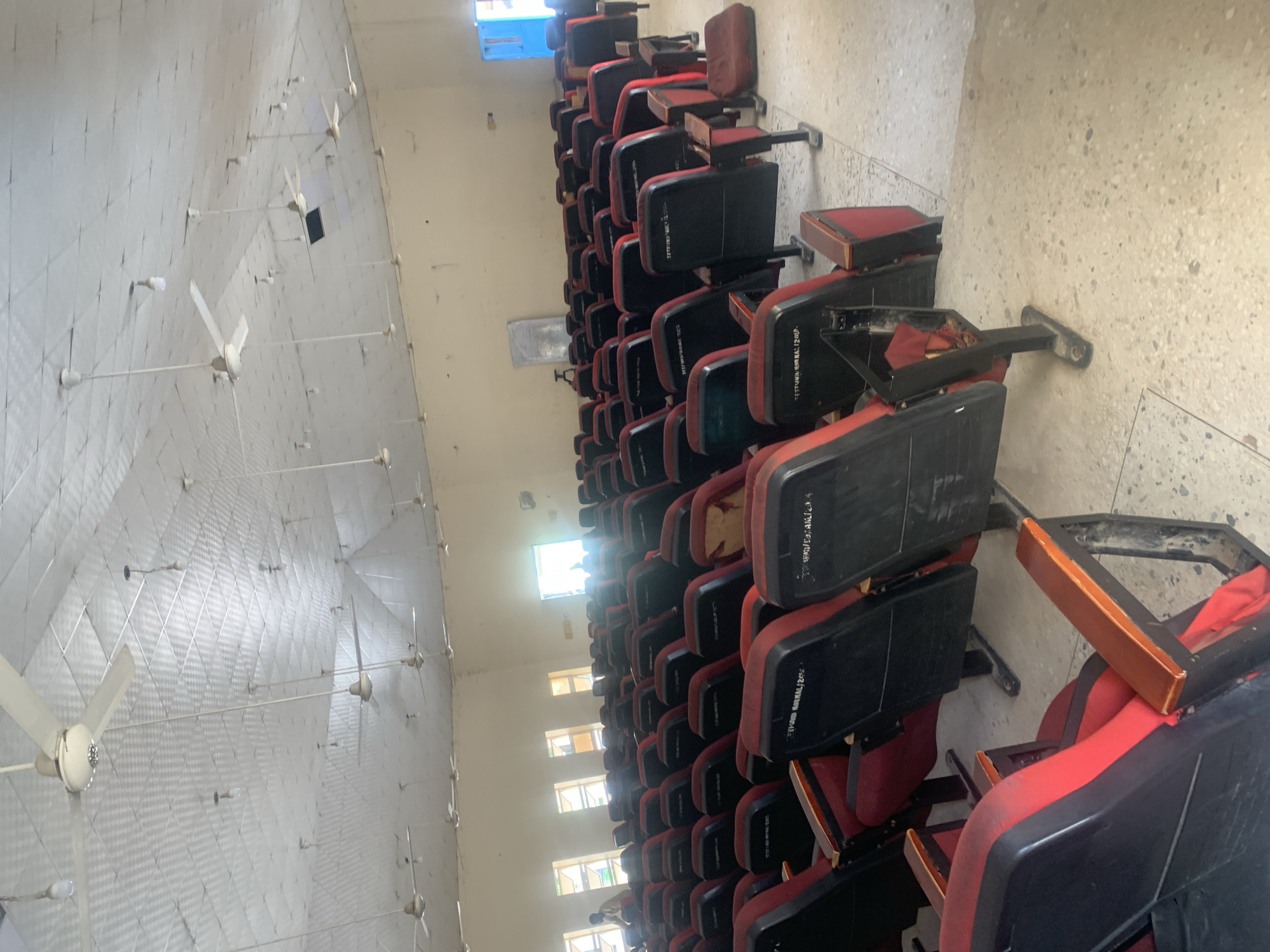 The image shows an interior view of a lecture hall or auditorium, likely located in Jalingo, Nigeria. Rows of empty, red and black fabric-covered seats with wooden armrests are arranged in a tiered fashion, facing a potential stage or presentation area, which is not fully visible. The ceiling is covered with white acoustic tiles and features numerous ceiling fans and bare light bulbs. Natural light streams in from windows on the left wall, and a brightly lit doorway is visible on the right side, revealing the silhouette of at least two individuals standing outside. The floor appears to be made of speckled concrete tiles. A few additional items like a small whiteboard on a stand and what looks like a cushion are visible on the floor. The overall impression is of a functional, though somewhat dated, academic or public gathering space.