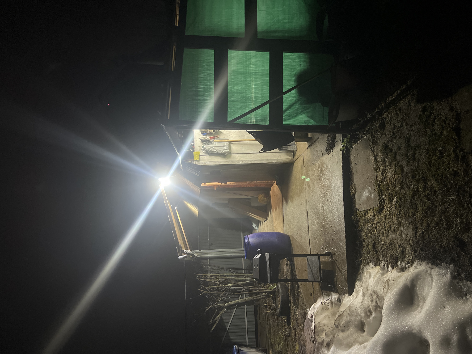 The image shows an outdoor scene at night. A bright light illuminates a covered area, likely a storage space or shed, with a green tarp covering the top and sides. Inside the shed, various objects are visible, including tools and containers. Outside the shed, there's a blue barrel, a small cart, and a pile of tree branches. Snow is present on the ground, suggesting a cold climate. The location is presumed to be in Chaikovsky, Russia, although there is no identifying information in the image itself to verify this. There are no people visible in the frame.