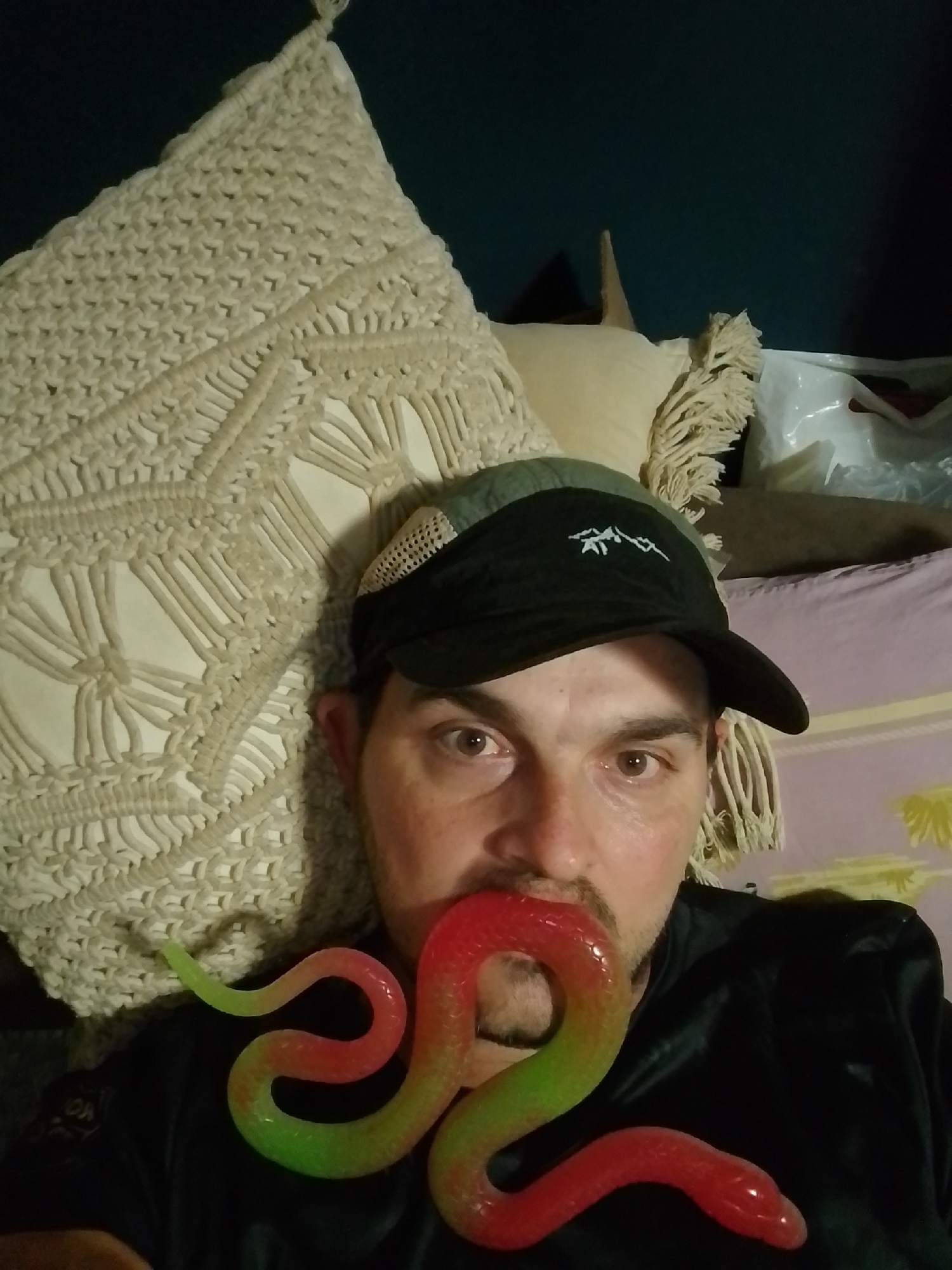A person wearing a dark baseball cap with an embroidered white mountain logo is lying down with their head resting on a beige macrame pillow. A colorful, coiled toy snake, transitioning from red to green, is placed in their mouth, extending outwards. The person's face is partially visible, showing their eyes, nose, and mustache. Behind them, other pillows and a glimpse of what appears to be a bag can be seen. The background is a dark wall. The image does not provide any information or context regarding the city of Wedel, Germany.