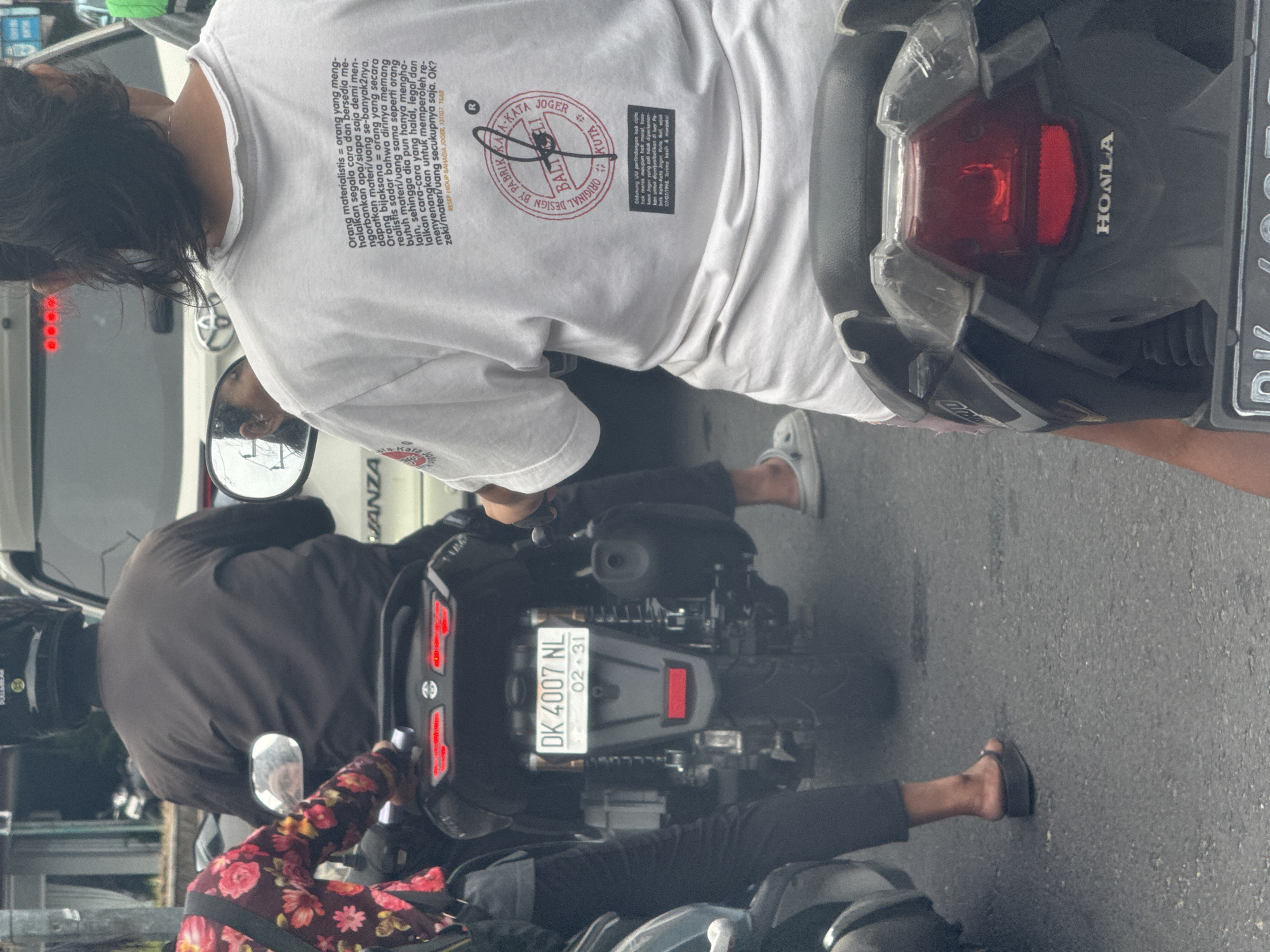 A medium-angle eye-level outdoor shot depicts a traffic scene on a street in Nusa Dua, Indonesia, featuring two motorbikes and a car. The primary focus is on the back of a person wearing a white t-shirt with Indonesian text and logos, riding a motorbike. The motorbike has a Honda logo and a license plate reading "DK 4007 NL" with an expiration date of "02-31". The person is wearing a white t-shirt with Indonesian text and logos. Another person riding a motorbike and wearing a dark brown top with a floral-patterned jacket is positioned in front of the first motorbike. A silver car, partially visible behind the first motorbike, has "AVANZA" written on it. The street scene includes parked vehicles in the background and street-level infrastructure. The lighting indicates a bright day, with the scene primarily in focus and accurately depicts various elements.