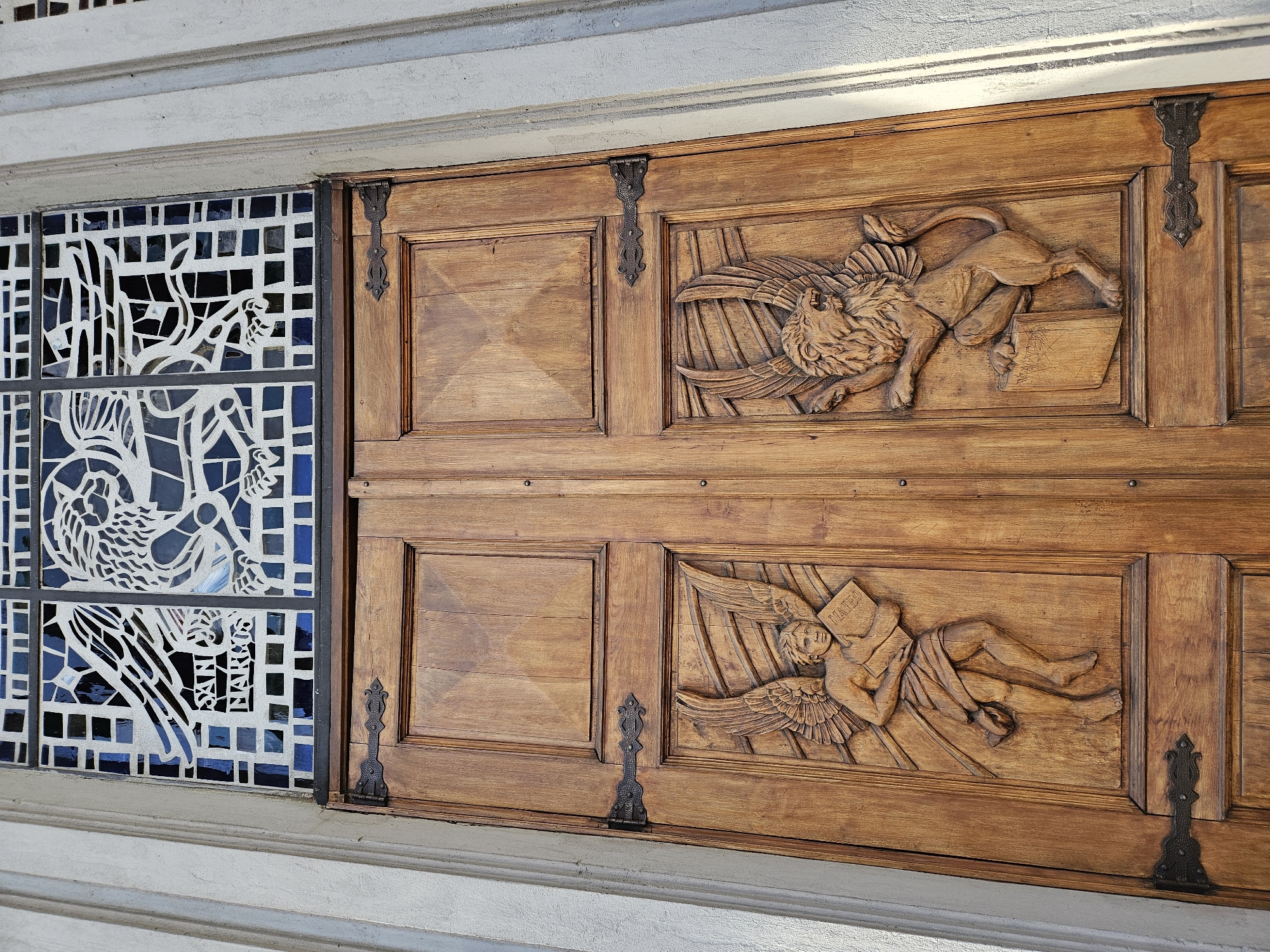 The image shows a section of a building's exterior, featuring a wooden door or panel adorned with carvings, alongside a modern stained-glass window.
The wooden structure comprises two large, rectangular panels, each featuring an intricate bas-relief carving. Both panels are secured with ornate, dark metal hinges. The upper carving depicts a powerful, winged lion, rendered with fine detail in its mane, musculature, and feathered wings. The lion appears to be roaring or bellowing, and its front paws rest upon an open book. The text on this book is indistinct. The lower carving presents a serene, winged human figure, possibly an angel or cherub, shown reclining. This figure is draped in cloth and holds an open book, clearly inscribed with the name "MATEO."
To the left of the wooden panels is a stained-glass window section. It features a contemporary, geometric design with a prominent, stylized bird-like figure in the lower portion. The glass is primarily white or clear, set against a mosaic of various blue hues. Within the design of this window, the words "SAN MARCOS" are visible, running vertically.
The scene is illuminated by bright daylight, casting subtle shadows and highlighting the textures of the rich brown wood, the carved details, and the varied surfaces of the stained glass. A light grey concrete or stone surface borders the architectural elements at the top and bottom. These symbolic carvings likely represent two of the four Evangelists: Saint Mark (winged lion, indicated by "SAN MARCOS" in the glass) and Saint Matthew (winged man, "MATEO" on the book). The setting is Osorno, Chile.