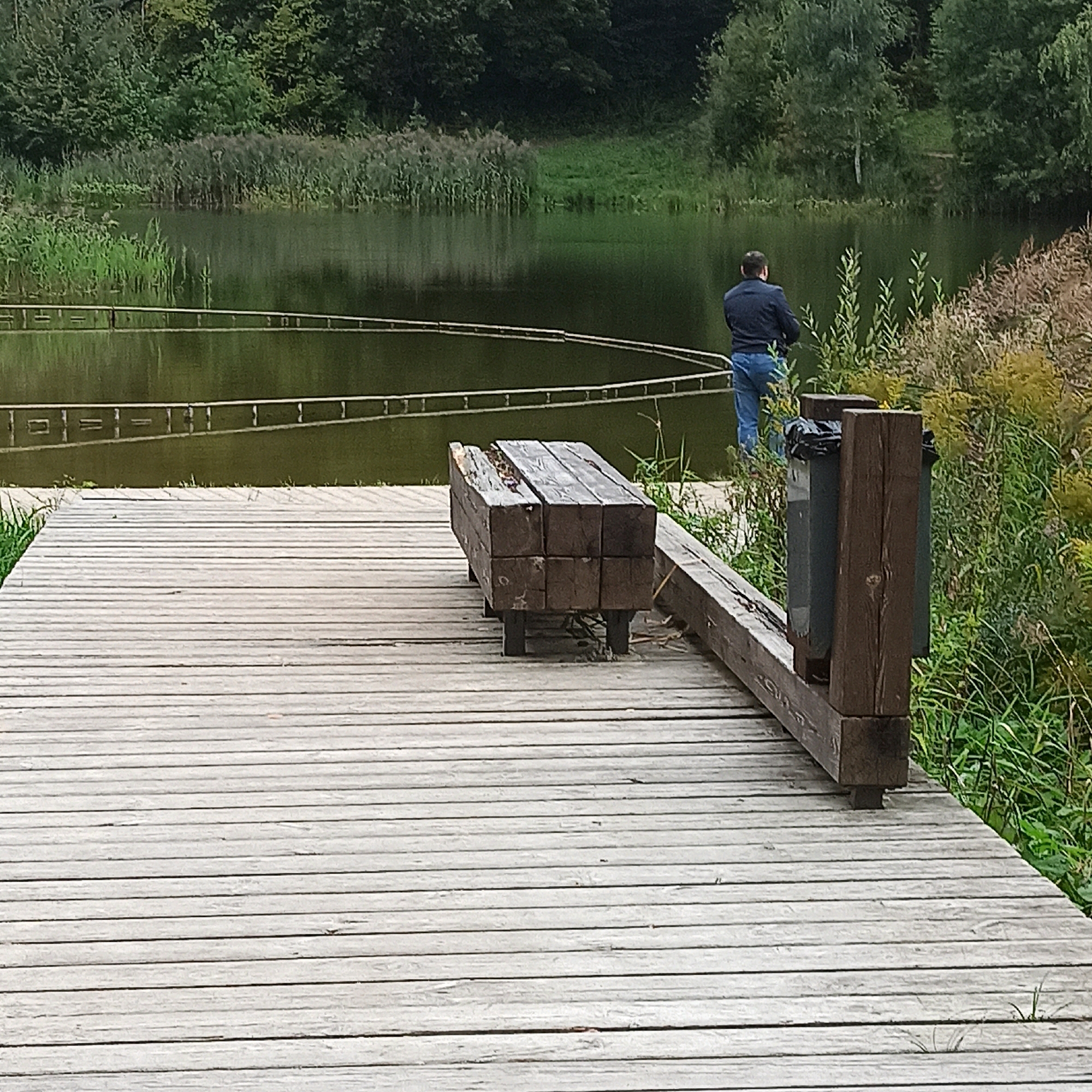 A person stands on a wooden dock, facing a lake. The person is wearing a dark jacket and blue jeans. In the foreground, there is a wooden bench and a trash can. The lake is surrounded by trees and reeds. The scene appears to be in a park or natural area, possibly in the city of Кузнечиха, Russia, given the provided context. The lighting suggests it is daytime, with the sun likely behind the viewer, casting shadows from the bench and dock.