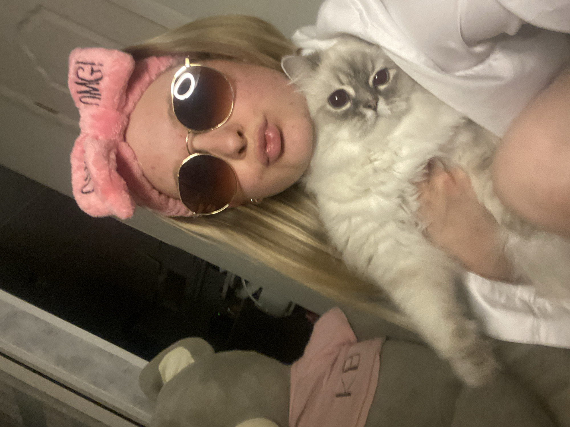The image captures a young individual, likely female, with blonde hair, centered in an indoor setting. The individual is wearing a fuzzy pink headband with the word "OMG!" printed in black text on the right side of the bow. They are also wearing gold-rimmed, round sunglasses with dark lenses, exhibiting a circular light reflection in the right lens. The individual's lips are pursed in a 'kiss face' expression. Held in their arms is a light-colored, long-haired cat, possibly a Ragdoll, with prominent dark eyes and darker points on its face. The cat is looking directly at the viewer. The individual is partially obscured by a white garment, possibly a robe or hoodie. To the left of the individual, a large grey plush toy, possibly a bear, is visible, wearing a pink t-shirt with the letters "KB" printed on it in black. The background to the left is dark and indistinct, featuring a vertical light-colored frame-like structure. The lighting is artificial and localized, contributing to reflections.