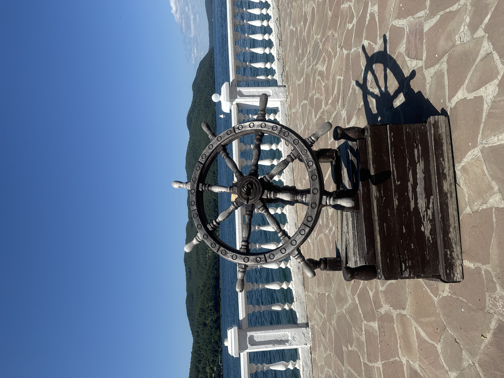 A wooden ship's wheel, mounted on a wooden base, is positioned on a paved walkway overlooking a large body of water. The walkway is made of irregular, flat stones in shades of tan and rust. A white, ornate railing runs along the edge of the walkway, separating it from the water. Behind the railing, the blue water stretches towards a distant, tree-covered shoreline. The sky is a clear, bright blue. The ship's wheel casts a distinct shadow on the paved ground. In the background, rolling hills covered in dark green trees line the opposite side of the water. The scene appears to be a scenic overlook in Listvyanka, Russia, a village on the southwestern shore of Lake Baikal. There are no people visible in the image.