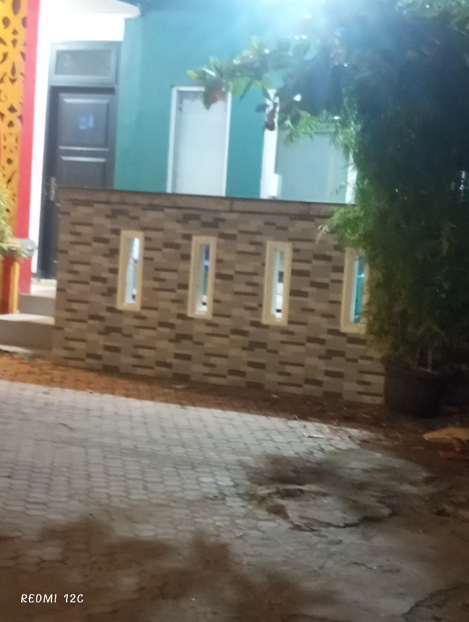 The scene is a residential area at night. A low brick wall with four vertical window slits stands in front of a green building. A tree in a pot is positioned on the right side of the wall. The ground appears to be paved with interlocking stones. A door with the number "24" is visible on the left side of the building. The image was taken with a Redmi 12C phone. The location is likely Lhokseumawe, Indonesia.