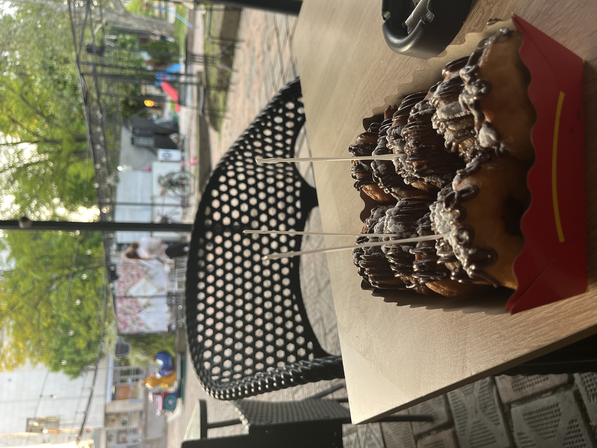 The image shows a partially eaten box of churros sitting on a light-colored wooden table outside. The churros are topped with chocolate and a white powdered substance, pierced with three white sticks for serving. A black, circular ashtray is partially visible on the table to the upper right. Behind the table is a black metal chair with a perforated seat and back. In the blurred background, there is a street scene with greenery, buildings, and people, suggestive of an urban environment. Polski Trambesh, Bulgaria, is not directly visually confirmed in the scene.