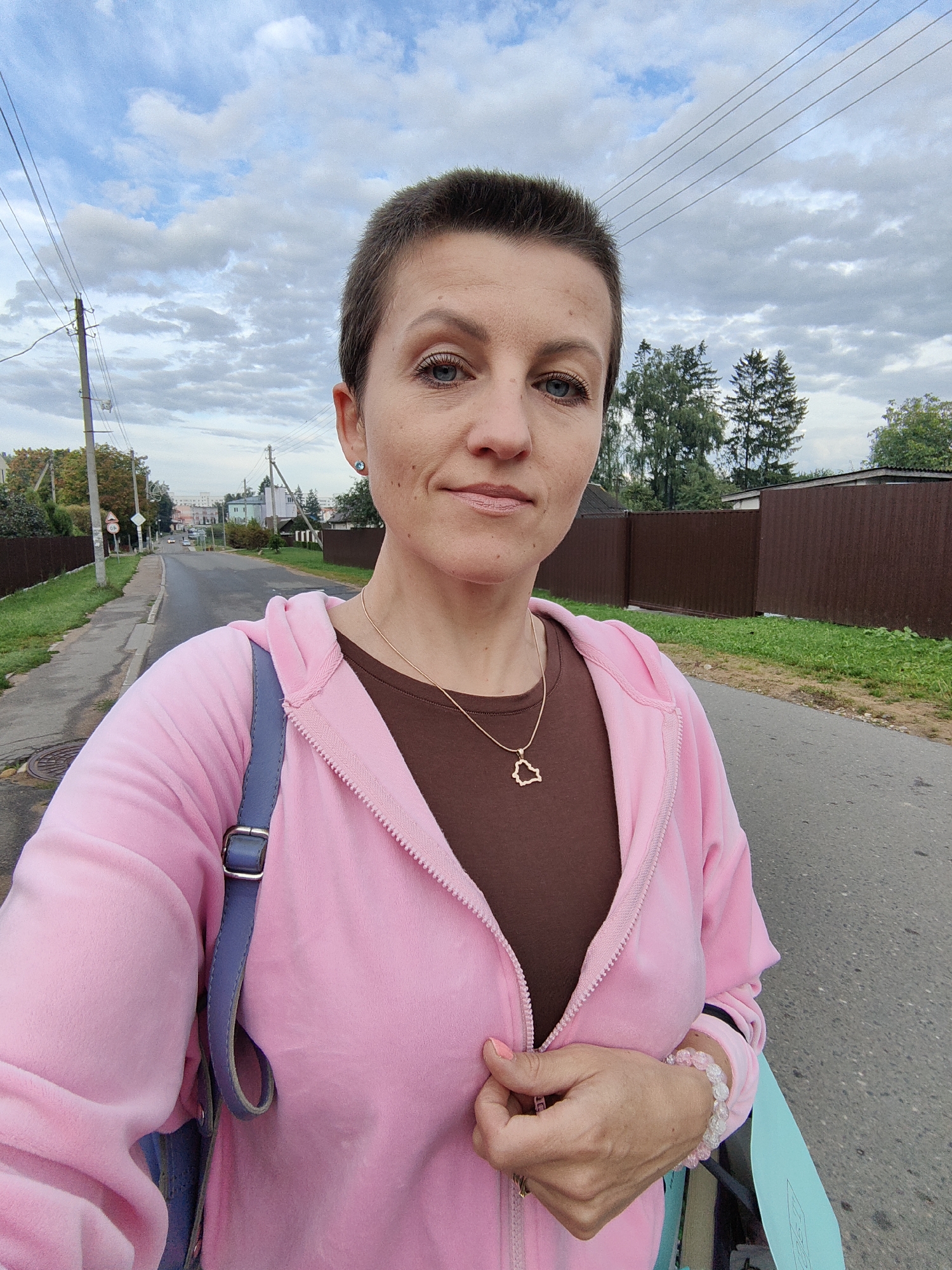 A woman with short brown hair and blue eyes is taking a selfie outdoors. She is wearing a pink velour zip-up hoodie over a brown t-shirt. She has a gold necklace with a heart-shaped pendant and a pink beaded bracelet on her left wrist. She is also wearing a blue strap from a bag across her chest. The background shows a paved road, a sidewalk, a grassy area with a brown fence, and trees. In the distance, there are houses and buildings, suggesting a suburban or semi-urban environment. The sky is cloudy. The location appears to be a street in Fanipal, Belarus, as evidenced by the presence of a traffic sign with Cyrillic lettering and the overall architecture.