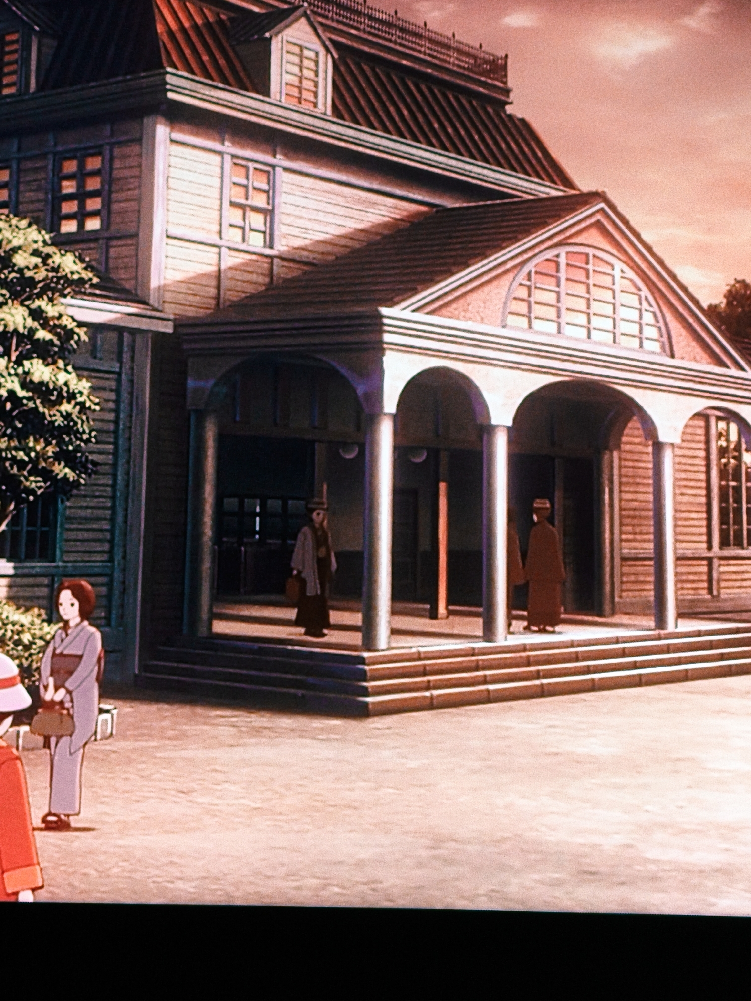 This is a digital illustration of a traditional Japanese-style building, possibly a school or temple, with a large wooden facade, a steeply pitched roof, and multiple windows. The building features an arched entrance supported by columns. Two figures, dressed in traditional Japanese attire, are visible standing on the steps of the building. A third figure, in a red outfit and a hat, is standing in the foreground to the left. The scene is set in what appears to be a courtyard or garden, with a tree to the left of the building. The sky is a warm, orange hue, suggesting either sunrise or sunset. The image does not depict any elements specific to Vladivostok, Russia, and therefore its location in that city cannot be determined from the visual information provided.