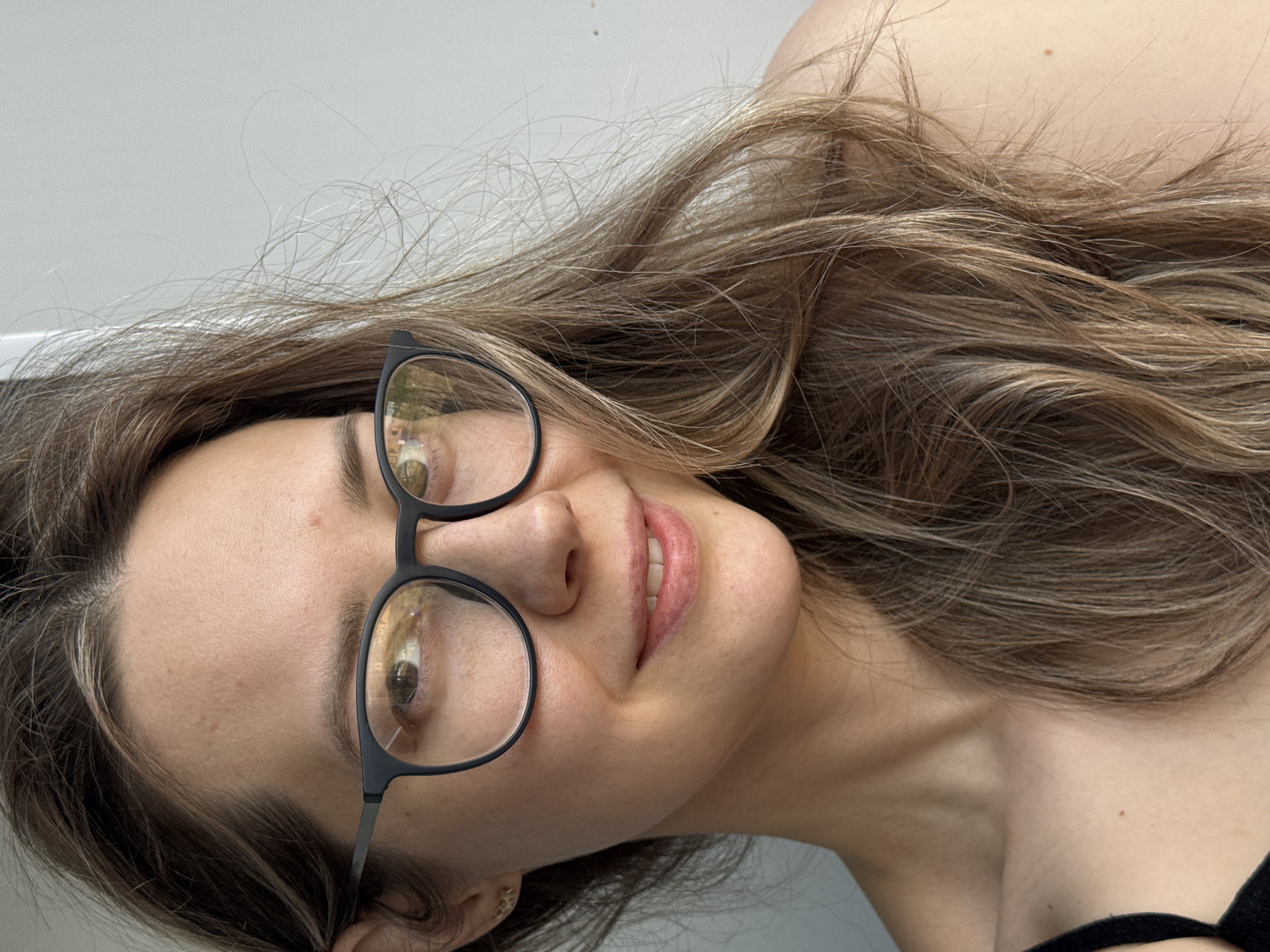 A woman with long brown hair and glasses is lying on a light-colored surface, possibly a bed or couch. She is smiling slightly. The location is unknown, and cannot be determined by the image.