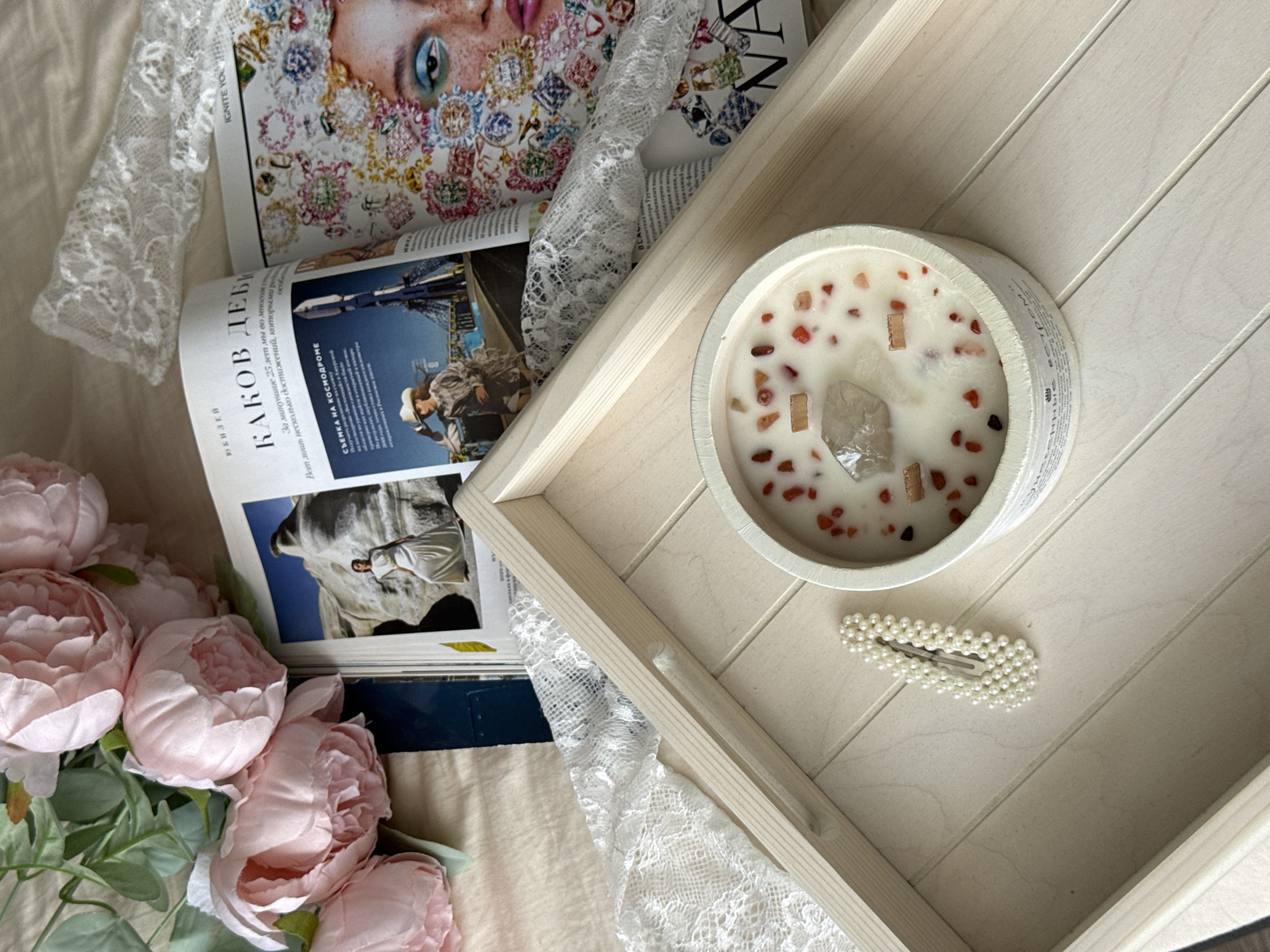 An overhead flat lay captures a serene indoor scene, likely set in Krasnodar, Russia, given the visible text. The central focus is a white, round candle with a wooden wick, embedded with small reddish-orange and clear crystal-like stones, resting on a light wooden tray with parallel ridges. Beside the candle, a white pearl-beaded hair clip lies on the tray.
Beneath the tray and on the left, several open magazines or books are visible. One features a close-up of a woman's face with elaborate, jeweled makeup, and another shows a man in a hat in an outdoor setting, and a person against a large rock formation. A piece of white lace fabric drapes elegantly over the magazines and across the scene. To the bottom left, a cluster of artificial light pink peonies with green leaves adds a touch of floral softness. The entire arrangement is set on a light-colored, soft fabric surface, likely a bedsheet.
Visible text on the magazines includes Russian words such as "ЮБИЛЕЙ" (Jubilee/Anniversary), "КАКОВ ДЕБИ" (How is Debi/Debbie), and "СЪЕМКА НА КОСМОДРОМЕ" (Filming at the Cosmodrome), along with fragments like "Вот лишь НОСАТЛЬКО достижений, которыми рей" (Here are just SOME achievements that are...) and "За минувание 25 лет мы во мноСтам ст" (For the past 25 years we are in many...). English text "IGNITE YOU" is also partially visible on one magazine. The scene evokes a sense of relaxation, personal care, and aesthetic pleasure.